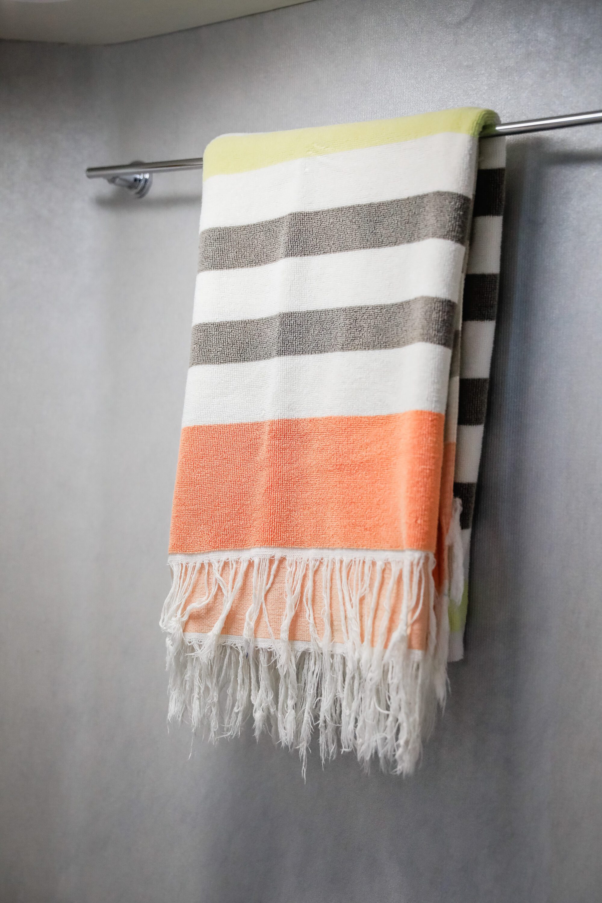 A striped towel with green, gray, white, and orange bands and white fringe hangs folded over a silver towel bar against a light-colored wall.