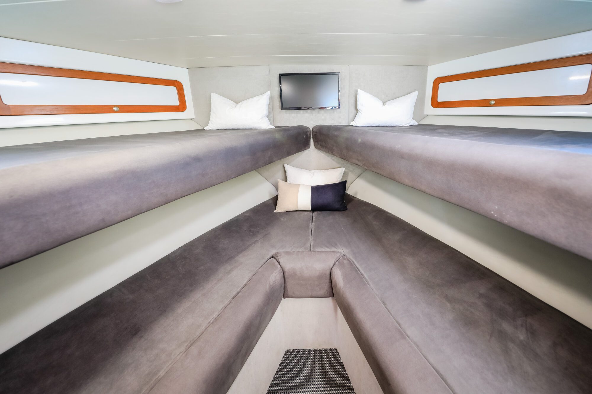 Interior of a boat cabin with two long cushioned benches on each side, a V-shaped cushioned seating area at the front, pillows, overhead storage compartments, a small TV mounted on the wall, and a rug on the floor.