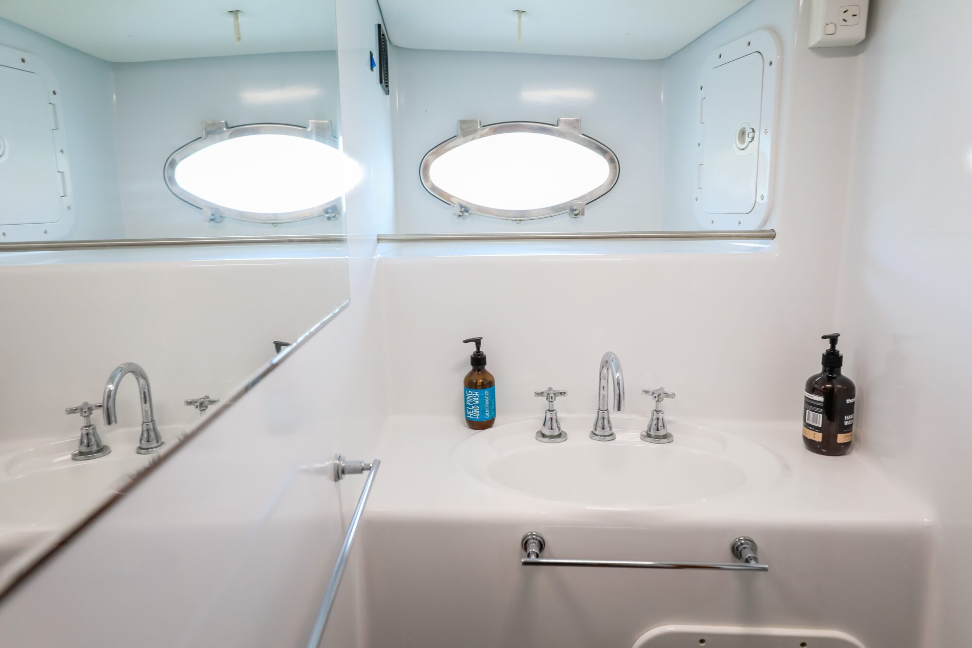 A white bathroom sink with two chrome faucets, two soap dispensers, a towel bar, and an oval-shaped window above, with part of the scene reflected in a mirror on the left.
