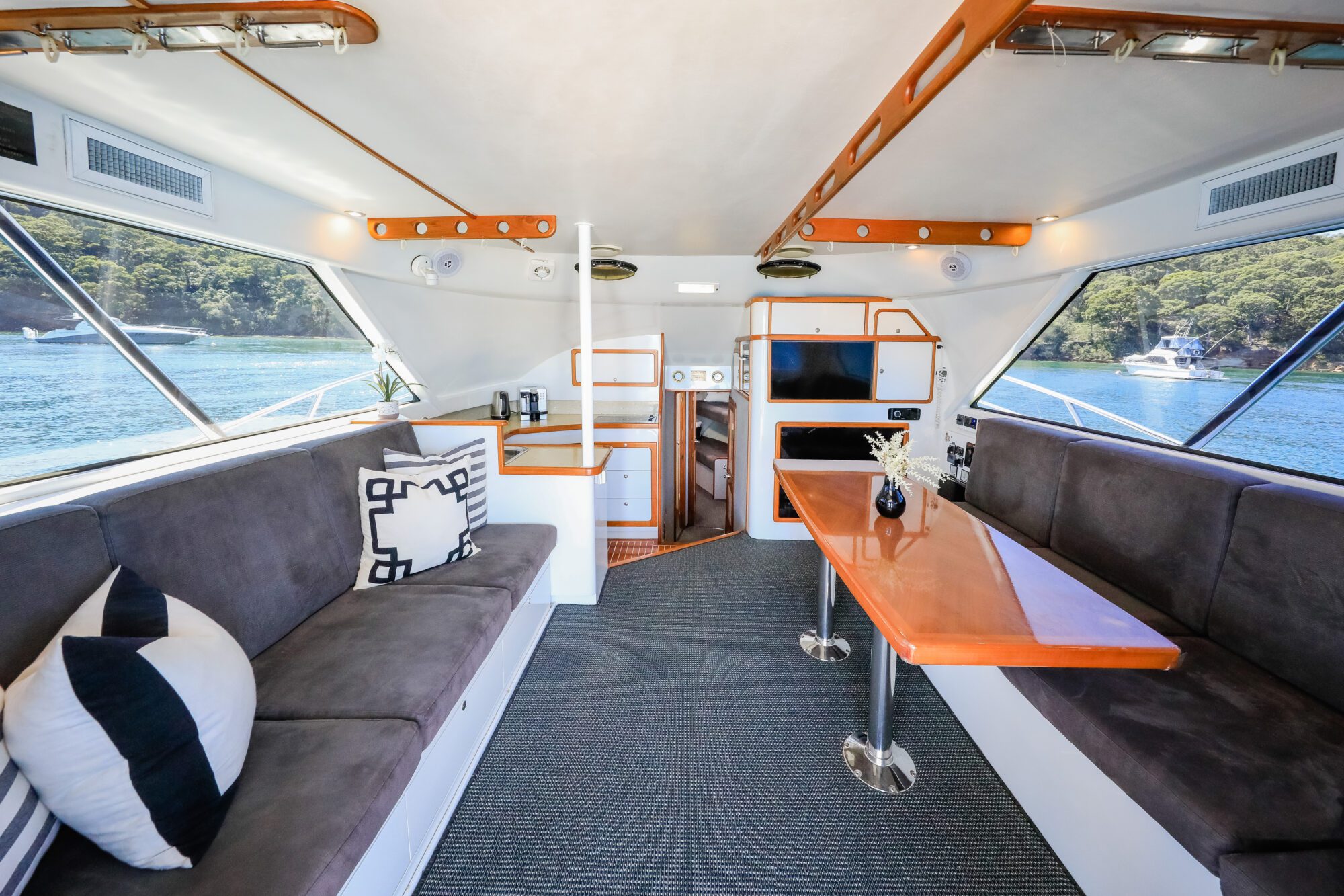 The interior of a modern boat with gray cushioned seating, a wooden table, large windows showing water and trees outside, and minimal decor including pillows and small plants.