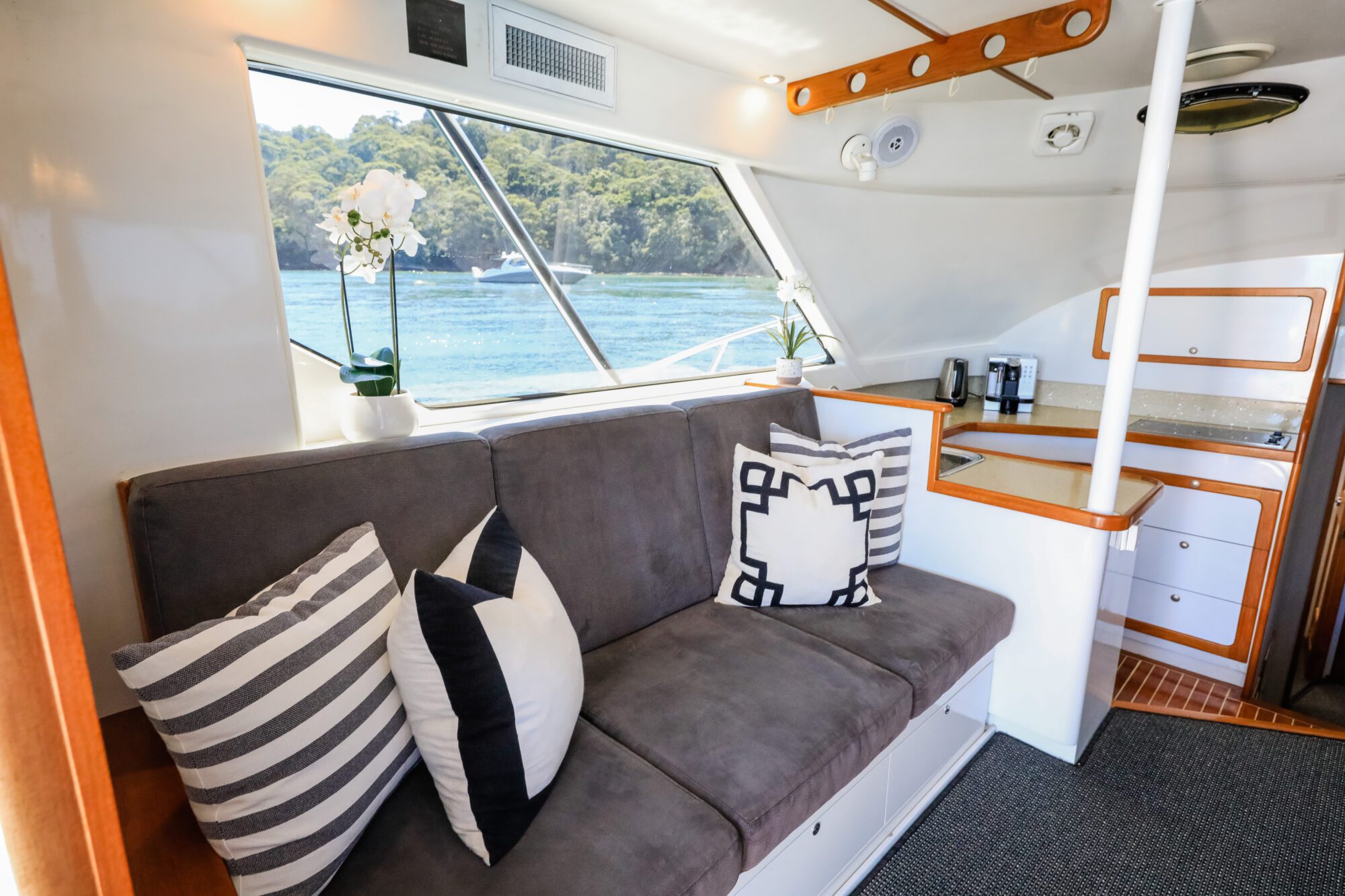 Interior of a boat with a gray cushioned sofa decorated with patterned pillows, a white orchid on the windowsill, and a kitchenette in the background; large windows show water and trees outside.