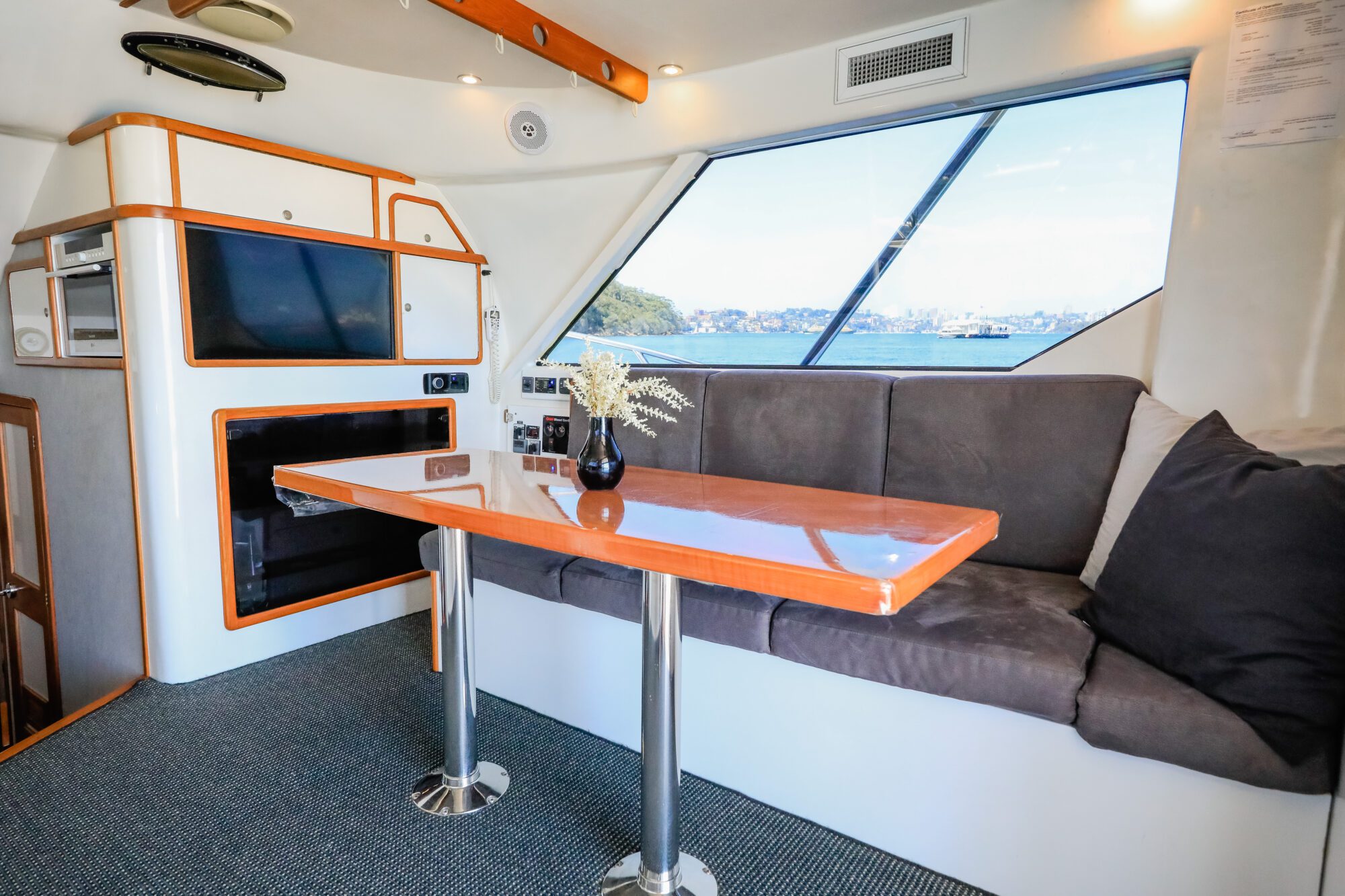 A modern yacht interior with a wooden table, gray cushioned seating, a TV, and large windows overlooking water and distant land. A vase with flowers sits on the table.