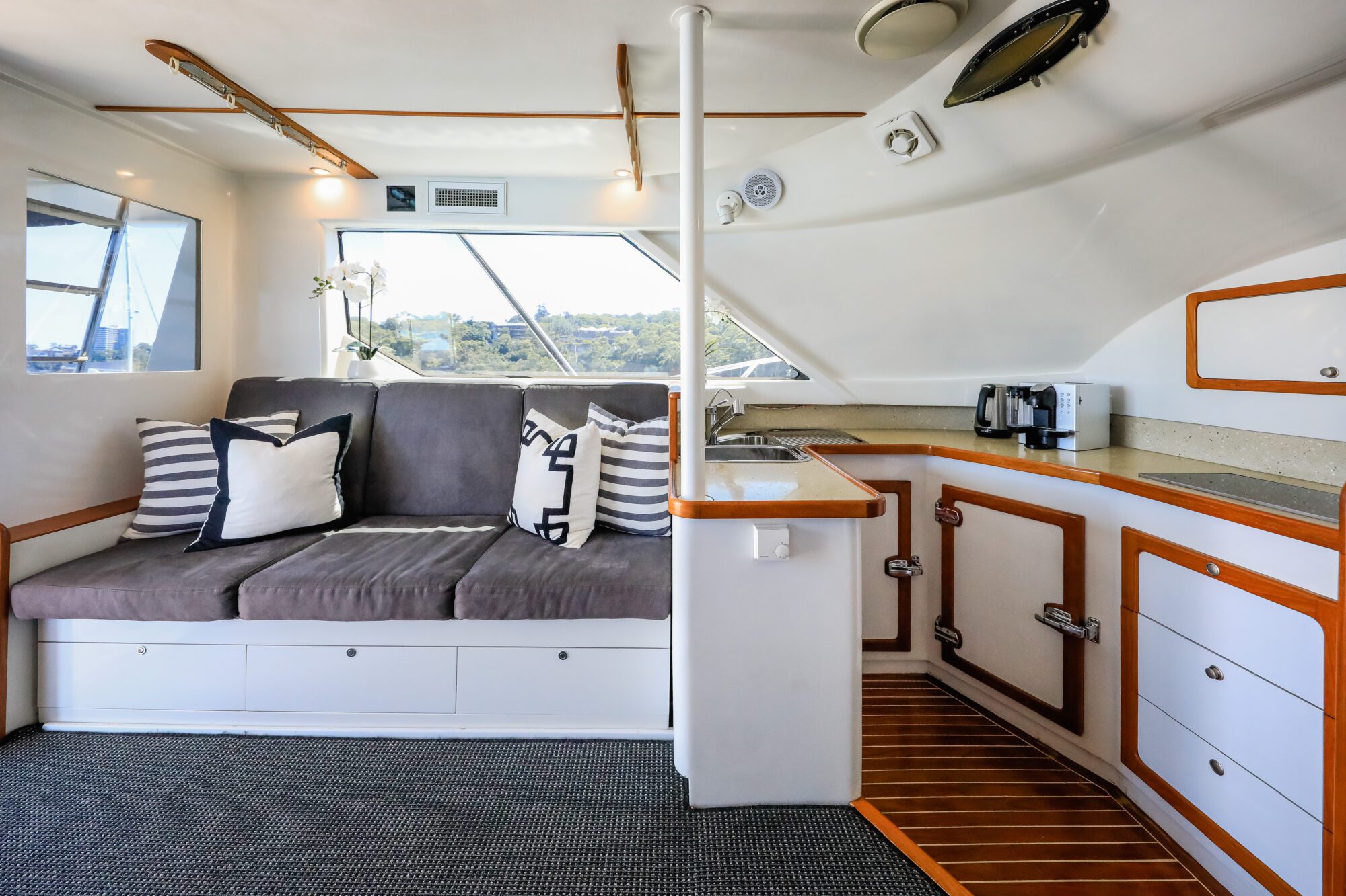 Modern yacht interior with a gray cushioned sofa, striped and patterned pillows, white cabinetry with wood trim, a small kitchenette with sink and coffee maker, large windows, and natural light filling the space.
