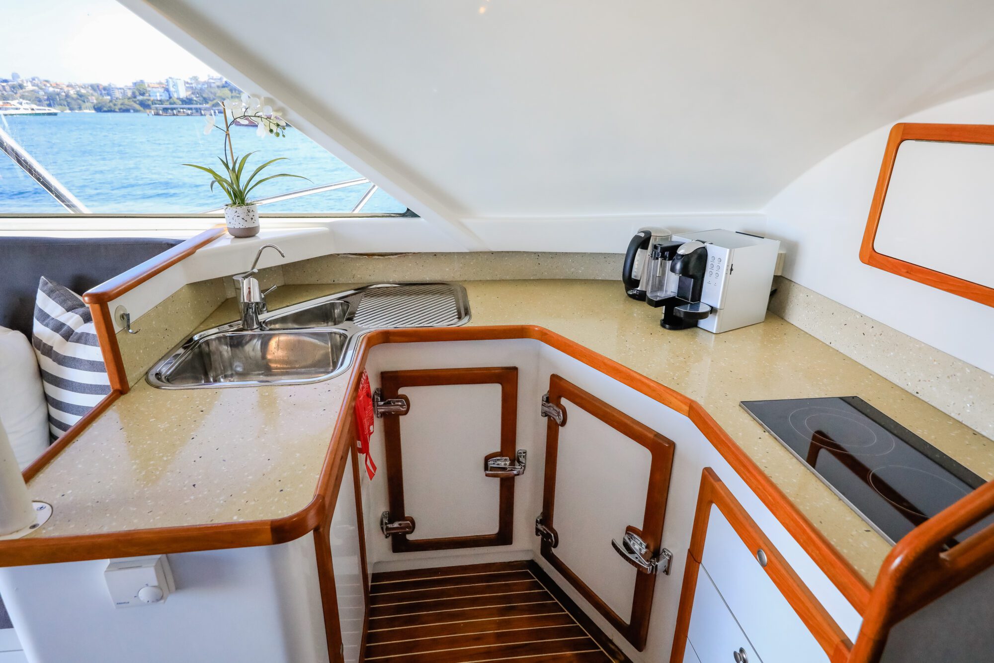 Modern yacht kitchen with a beige countertop, sink, stove, coffee maker, and white cabinets with wood trim. Window provides view of water and shoreline. White orchid sits on the counter.