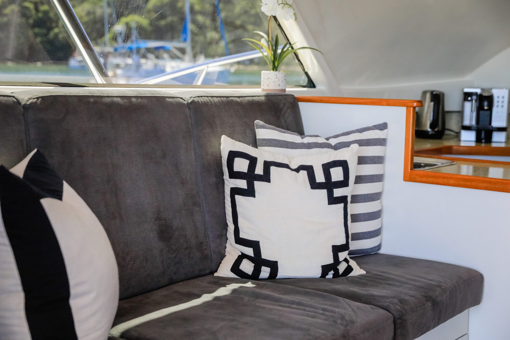 A close-up of a grey couch with decorative black, white, and striped pillows on a boat. In the background, a potted plant, kitchen counter, coffee maker, and blurred view of water and trees are visible through the window.