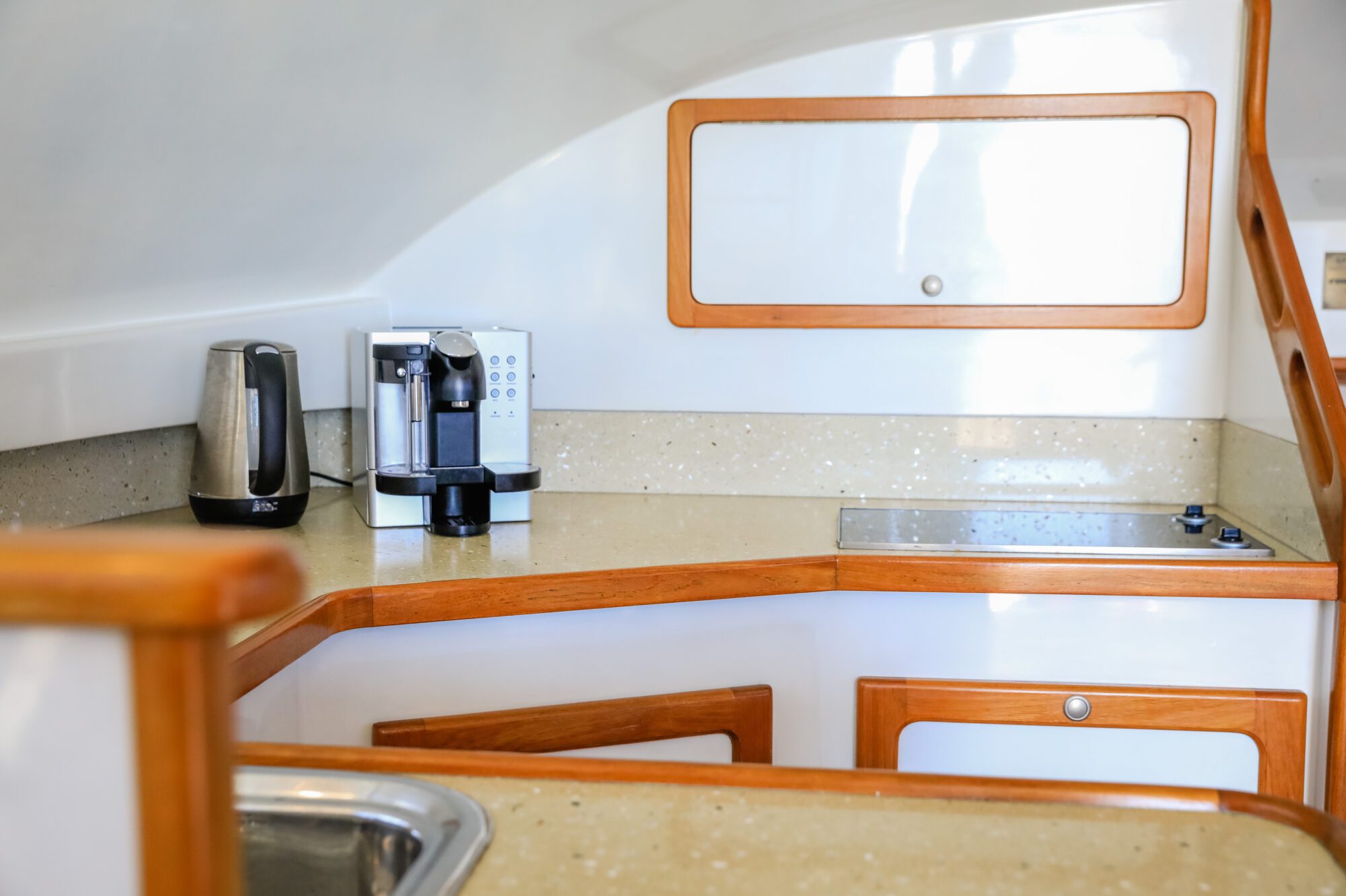 A compact kitchen with beige countertops, wooden trim, a sink, an electric kettle, a coffee machine, a stove with two burners, and white cabinets with wooden frames. The area is well-lit and clean.