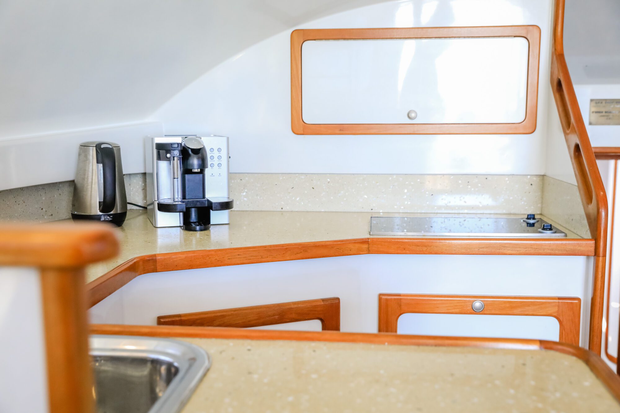A small modern kitchen with light countertops, a silver kettle, a coffee maker, a built-in electric stove, a sink, and wooden trim around the cabinets and counter edges.