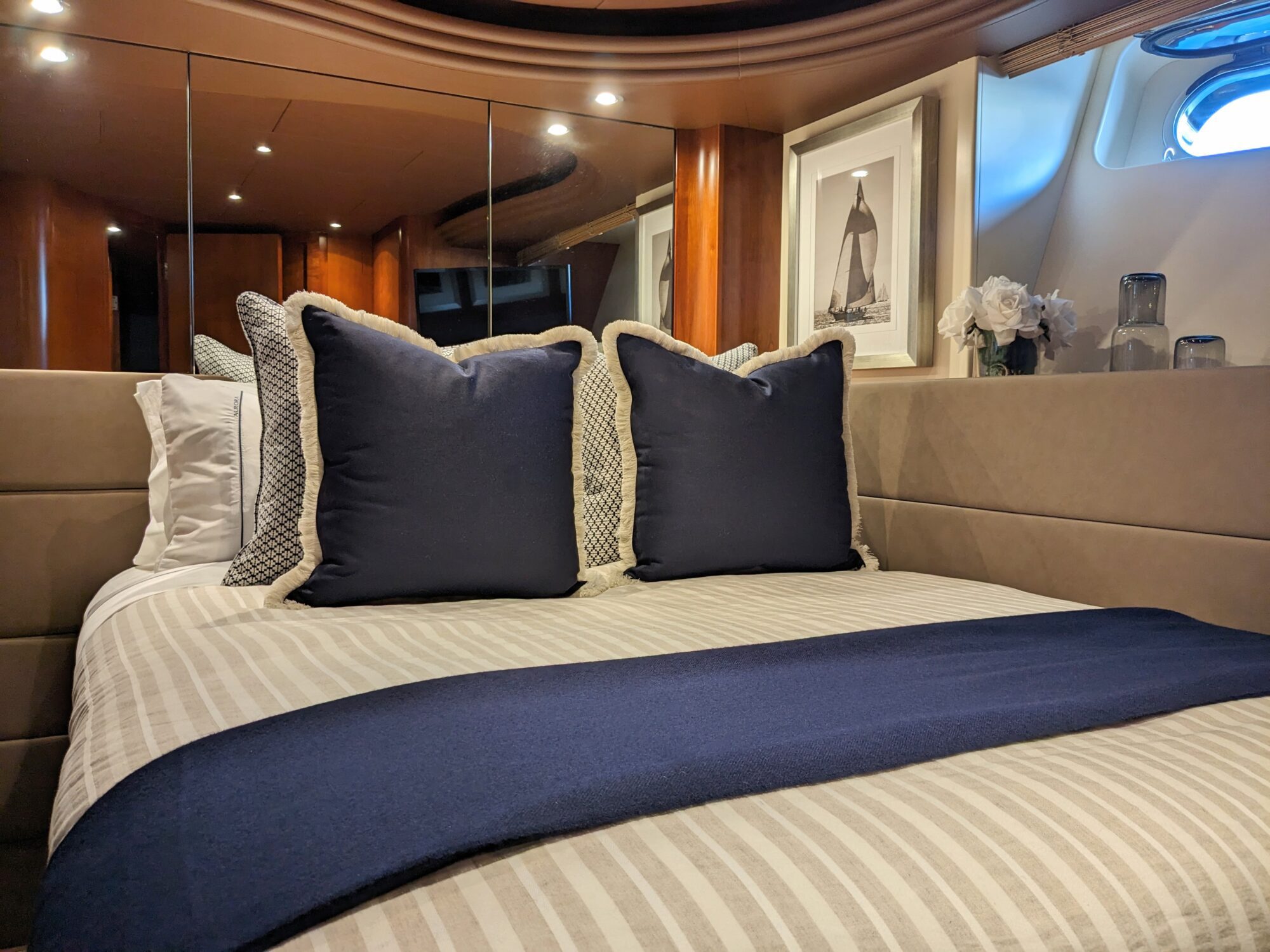 A neatly made bed with striped bedding, blue decorative pillows, and a blue throw in a modern, wood-paneled room with framed nautical art, a small vase of white flowers, and a porthole letting in light.