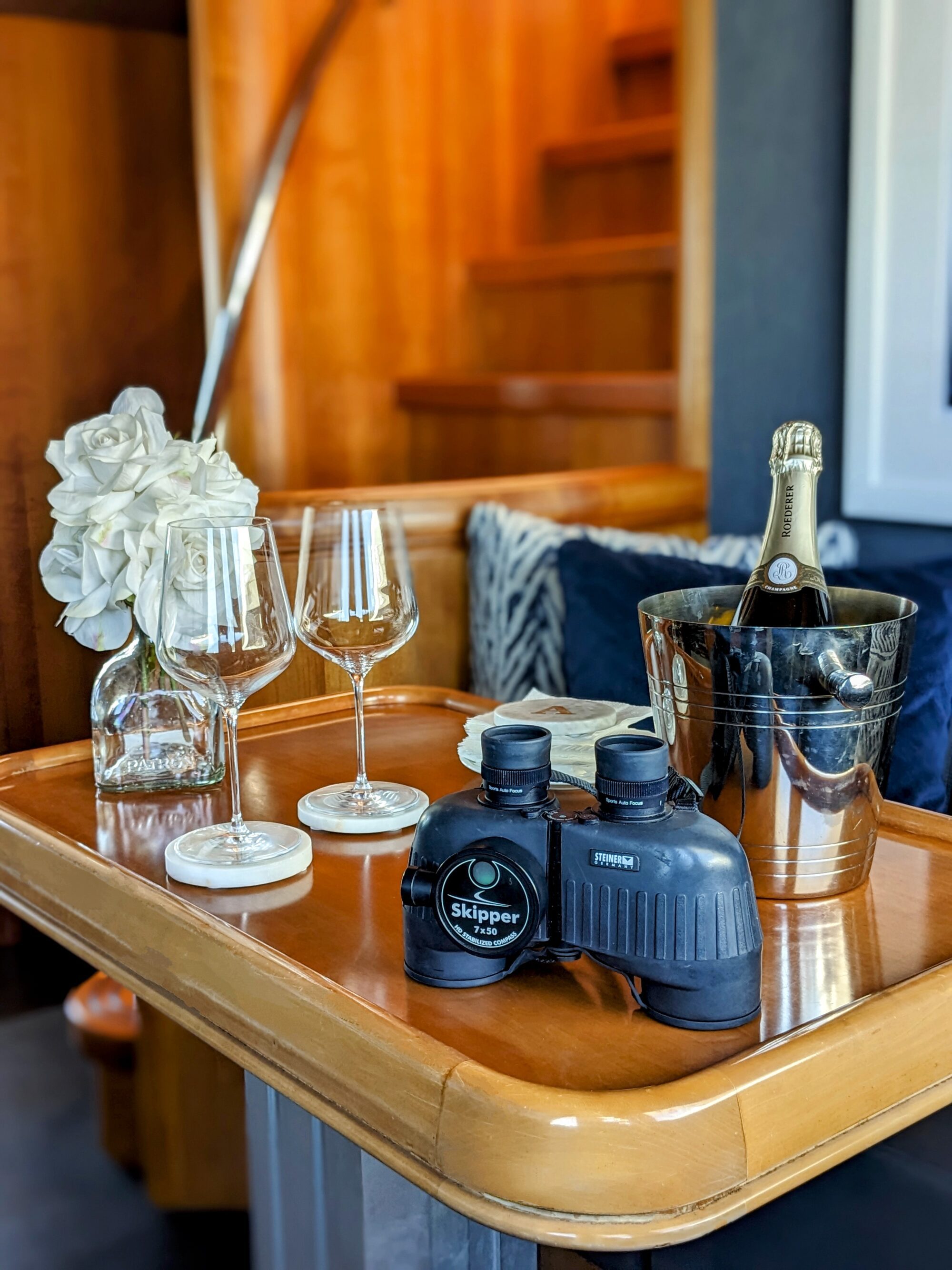 A wooden table holds two wine glasses, a bottle of champagne in an ice bucket, white roses in a vase, coasters, and binoculars. A staircase with wooden steps is in the background.