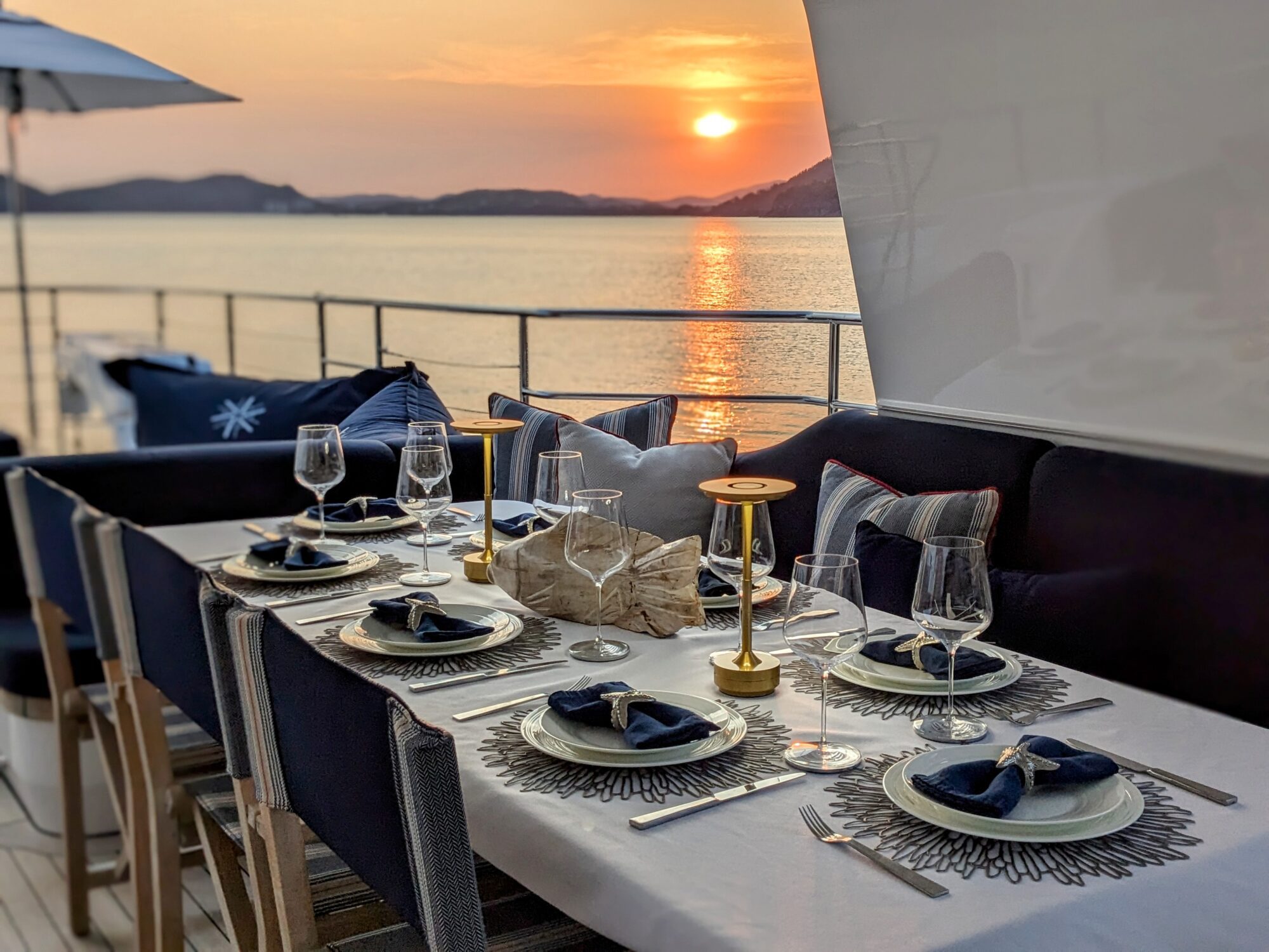 A beautifully set dining table on a yacht deck overlooks a calm sea at sunset. The table has elegant plates, navy napkins, wine glasses, and a golden candleholder, with plush seating around.