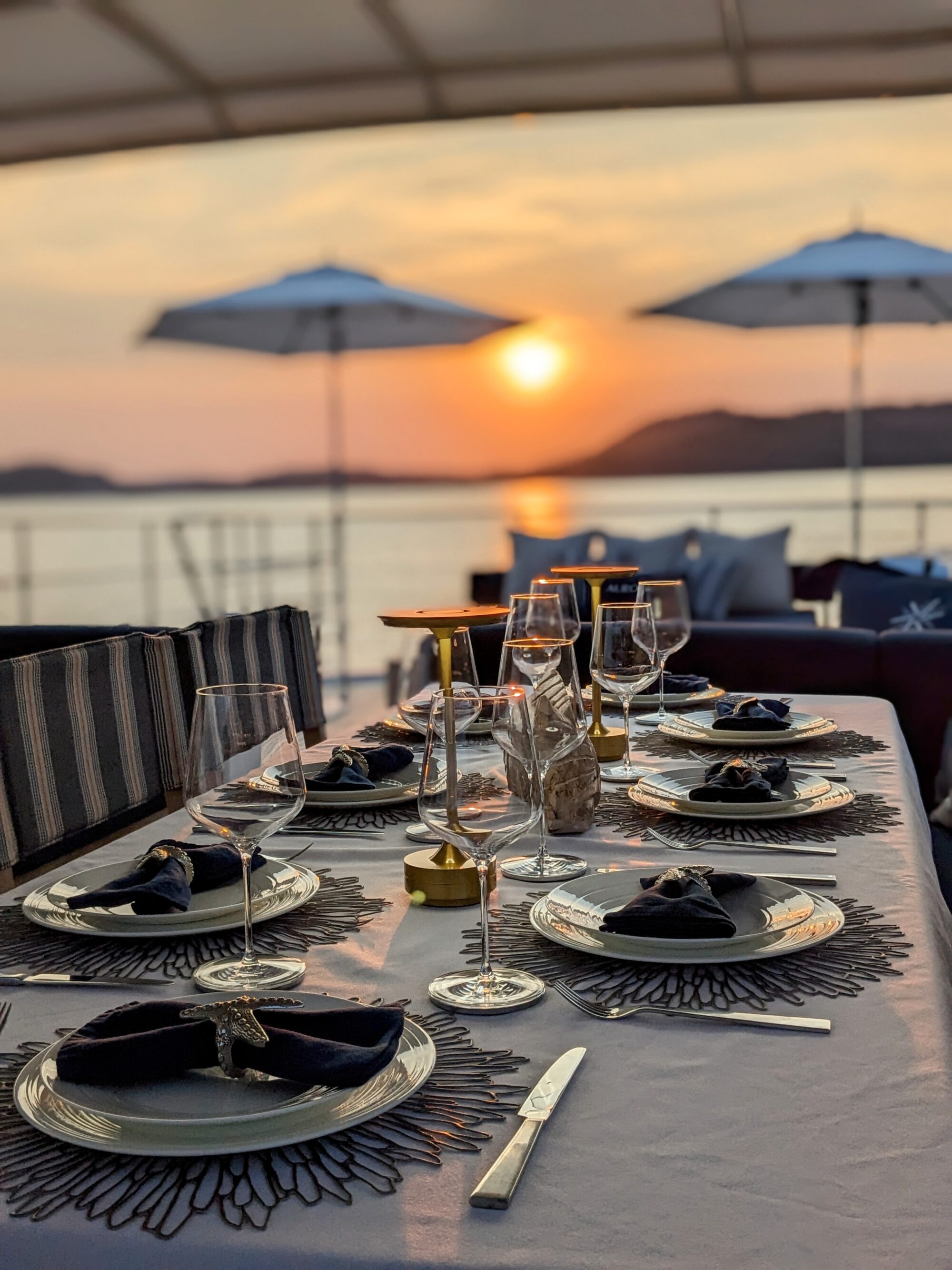 Elegant outdoor dining table set with plates, wine glasses, and navy napkins, overlooks a calm sea at sunset. Two white umbrellas and distant hills are visible under a warm orange sky.