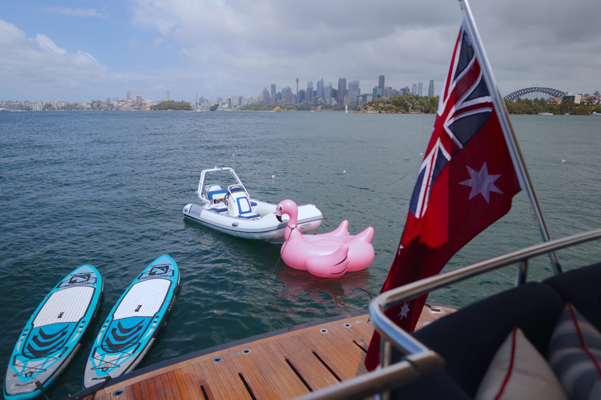 A boat deck with two paddleboards, an inflatable boat, and a pink flamingo float on the water. An Australian flag is visible, with a city skyline and Sydney Harbour Bridge in the background.