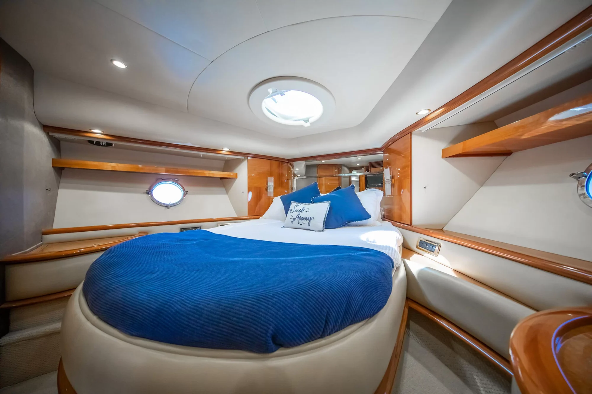 A cozy boat cabin with a round bed covered in blue and white bedding, wood accents, shelves, and round windows letting in natural light.