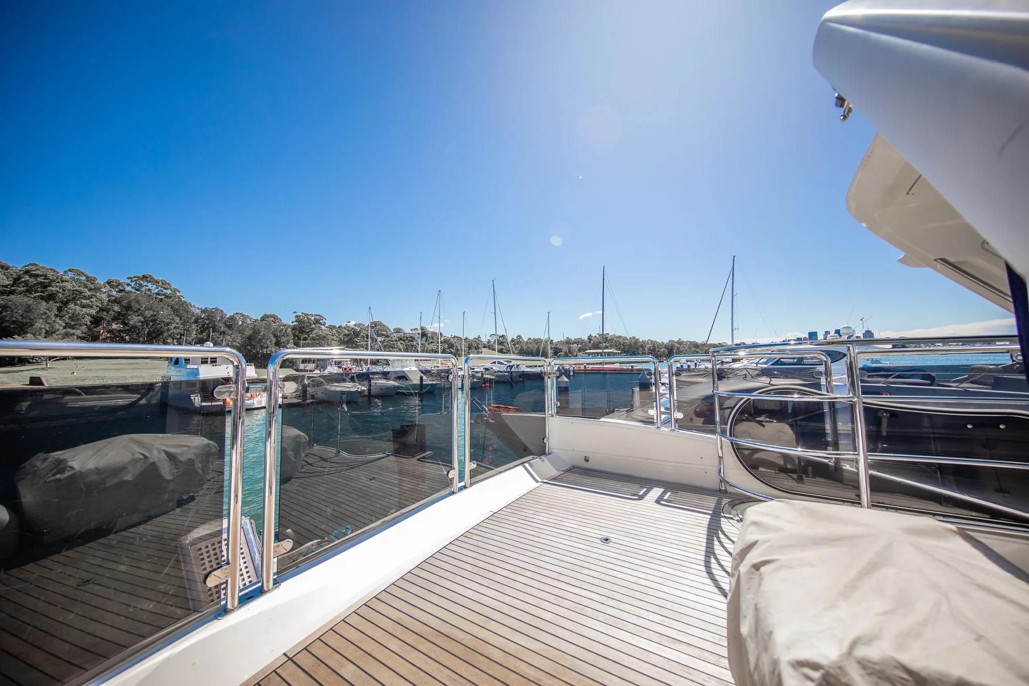 View from the deck of a yacht overlooking a marina with several boats and sailboats docked, surrounded by calm water and trees under a clear blue sky.