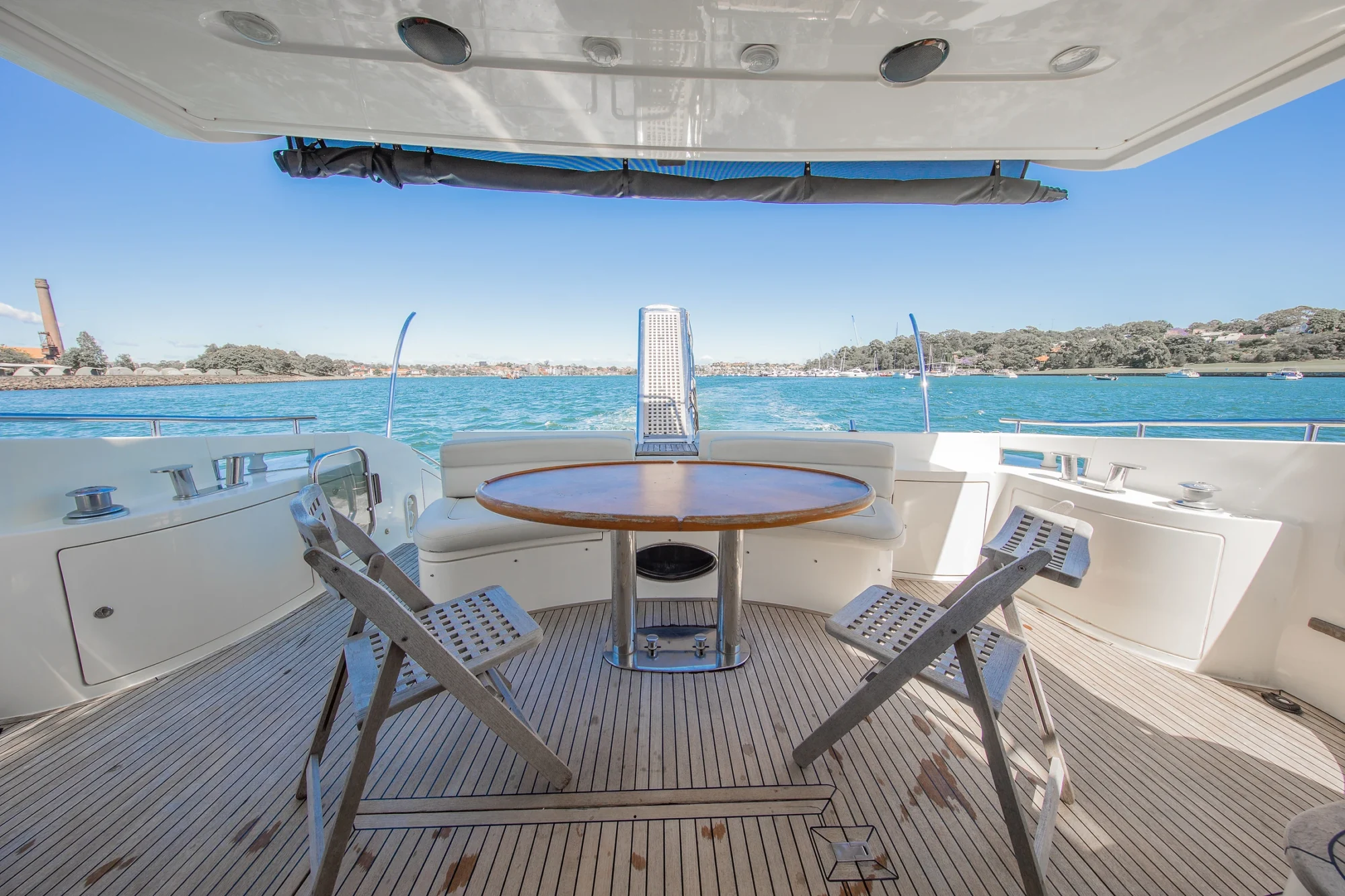 View from the deck of a yacht showing a round wooden table with four folding chairs, overlooking blue water and distant green shoreline under a clear sky.