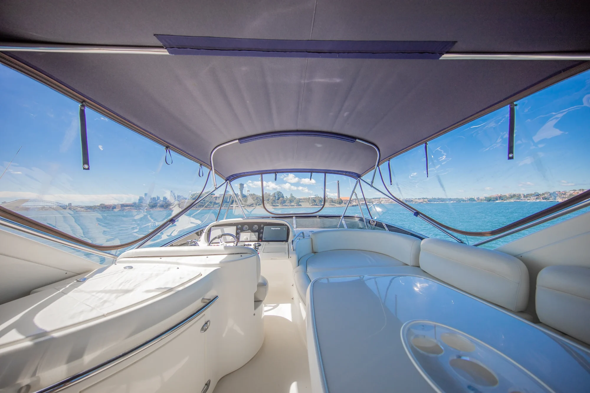 The image shows the interior of a modern yacht with white seating, a steering console, and large windows offering a clear view of the blue ocean and sky. The space is covered by a canopy, creating a shaded, relaxed atmosphere.