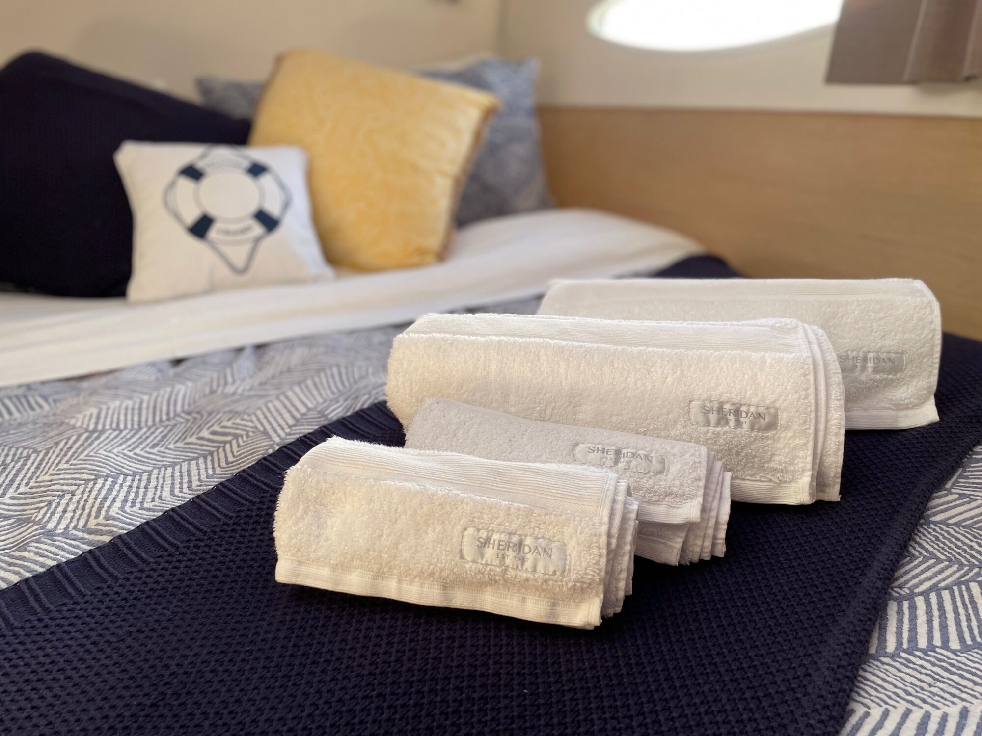 Four neatly rolled white towels are placed on a bed with navy and white bedding. There are decorative pillows in the background, including one with a life preserver design. Sunlight filters in through a window.