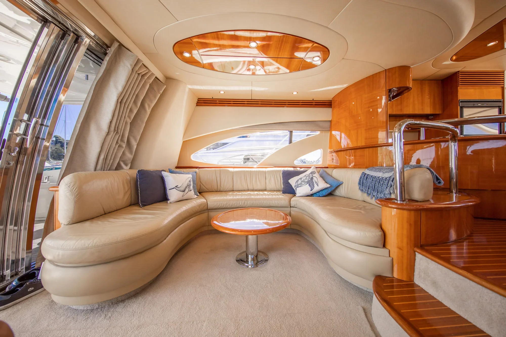 Luxurious yacht interior featuring a curved beige leather sofa with nautical-themed pillows, a round wooden coffee table, large windows, polished wood accents, and a staircase with metal railing.