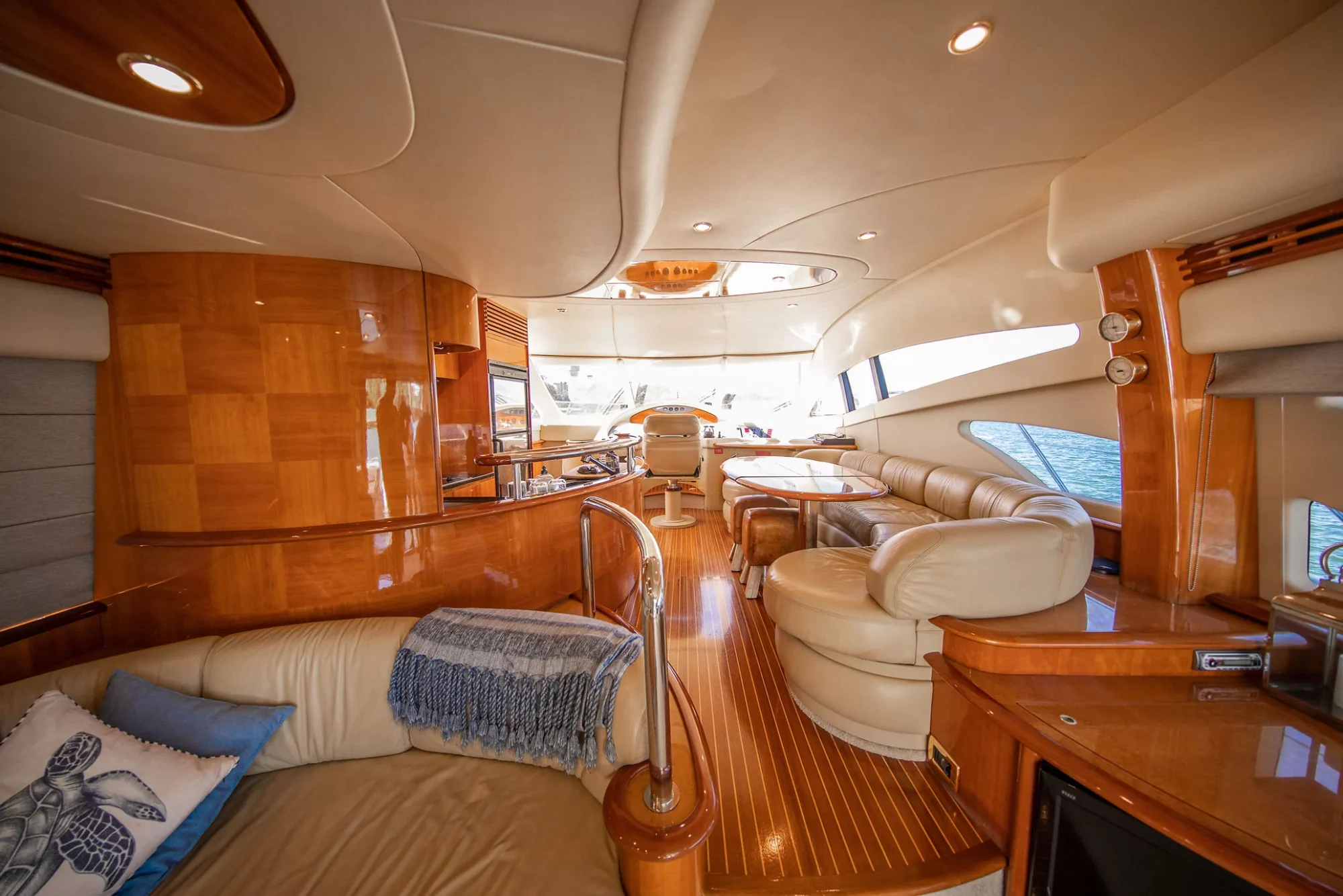 The image shows the luxurious interior of a yacht with cream leather sofas, wood floors, polished wood paneling, a dining area, and large windows overlooking the water, creating a bright and elegant atmosphere.