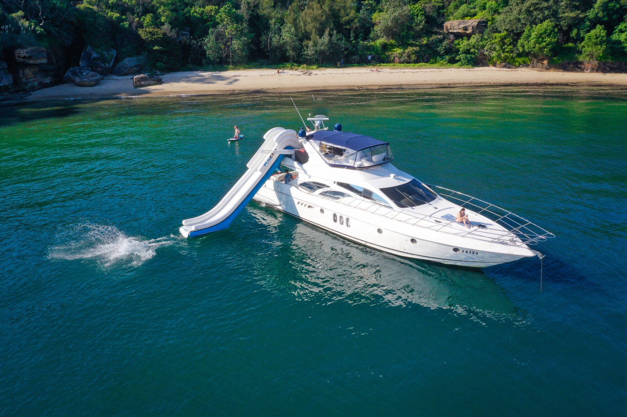 A white yacht anchored near a sandy beach with lush greenery. A person is sliding down a blue and white water slide from the yacht, while another person stands on the deck and someone is paddleboarding nearby.