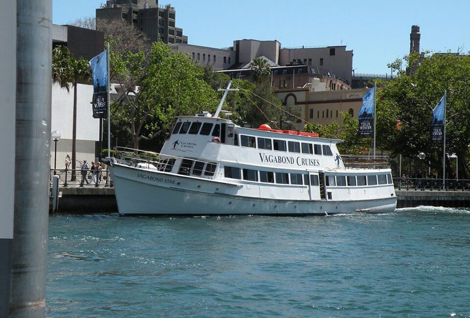 A white cruise boat named "Vagabond Star" from Vagabond Cruises is docked by a pier next to a tree-lined waterfront with buildings in the background and blue water in the foreground.