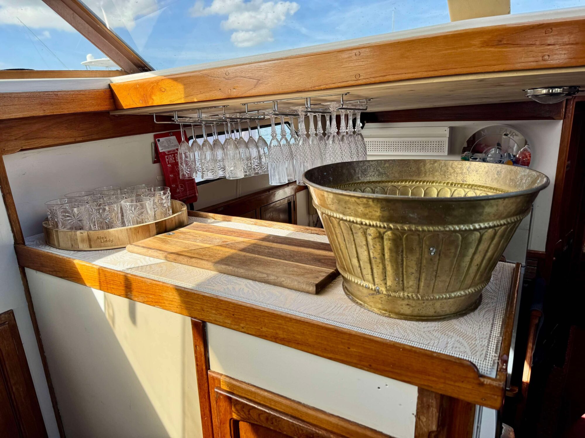 The Mayfair brass drink tub rests on a wooden counter beside a round tray with glasses and two cutting boards. Above, hanging wine glasses are stored, while sunlight pours in through the window.