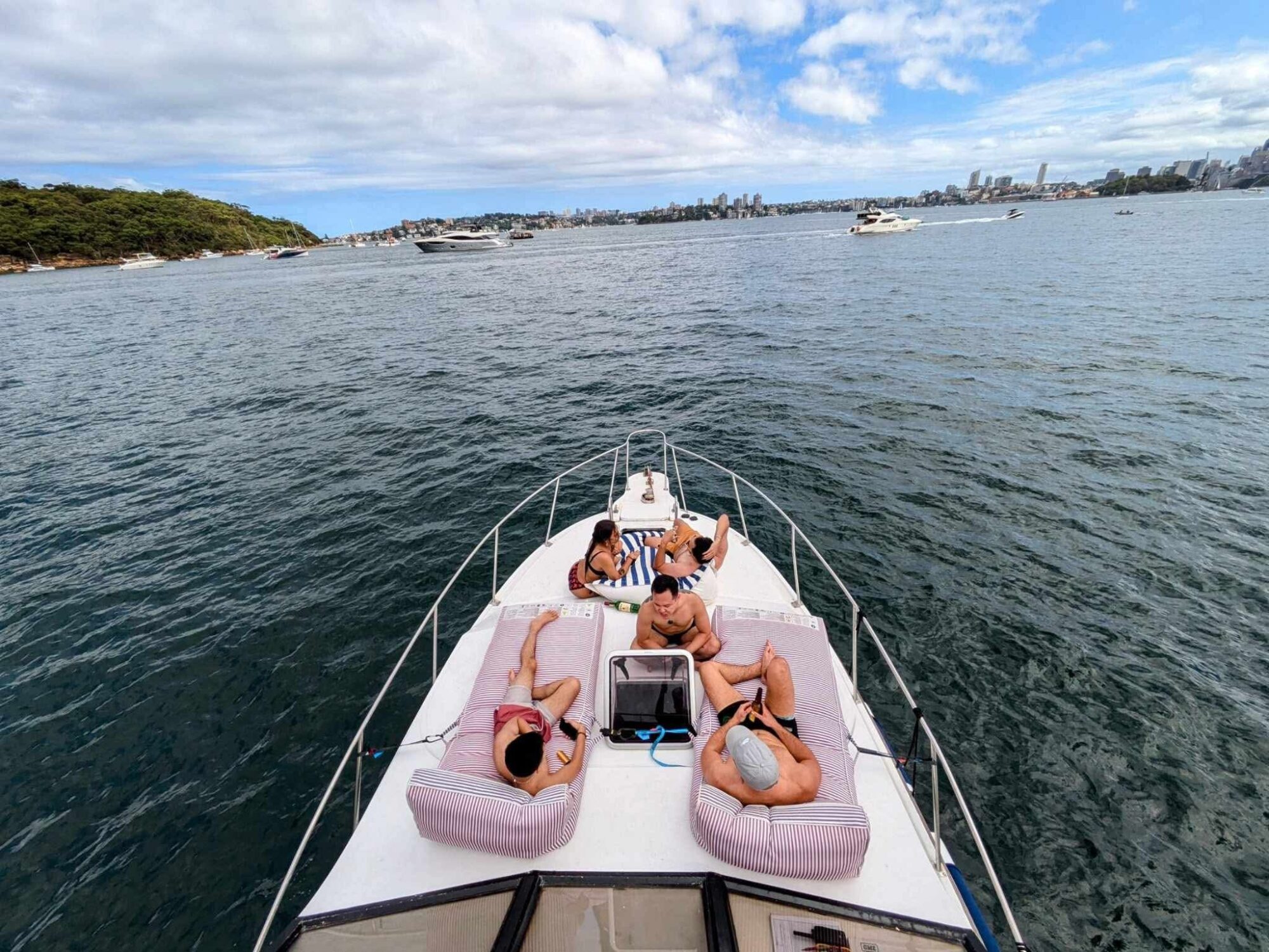 Four people relax on the bow of the Mayfair yacht with pink cushions, enjoying a sunny day on calm water. The shoreline, city skyline, and other boats are visible under a partly cloudy sky.
