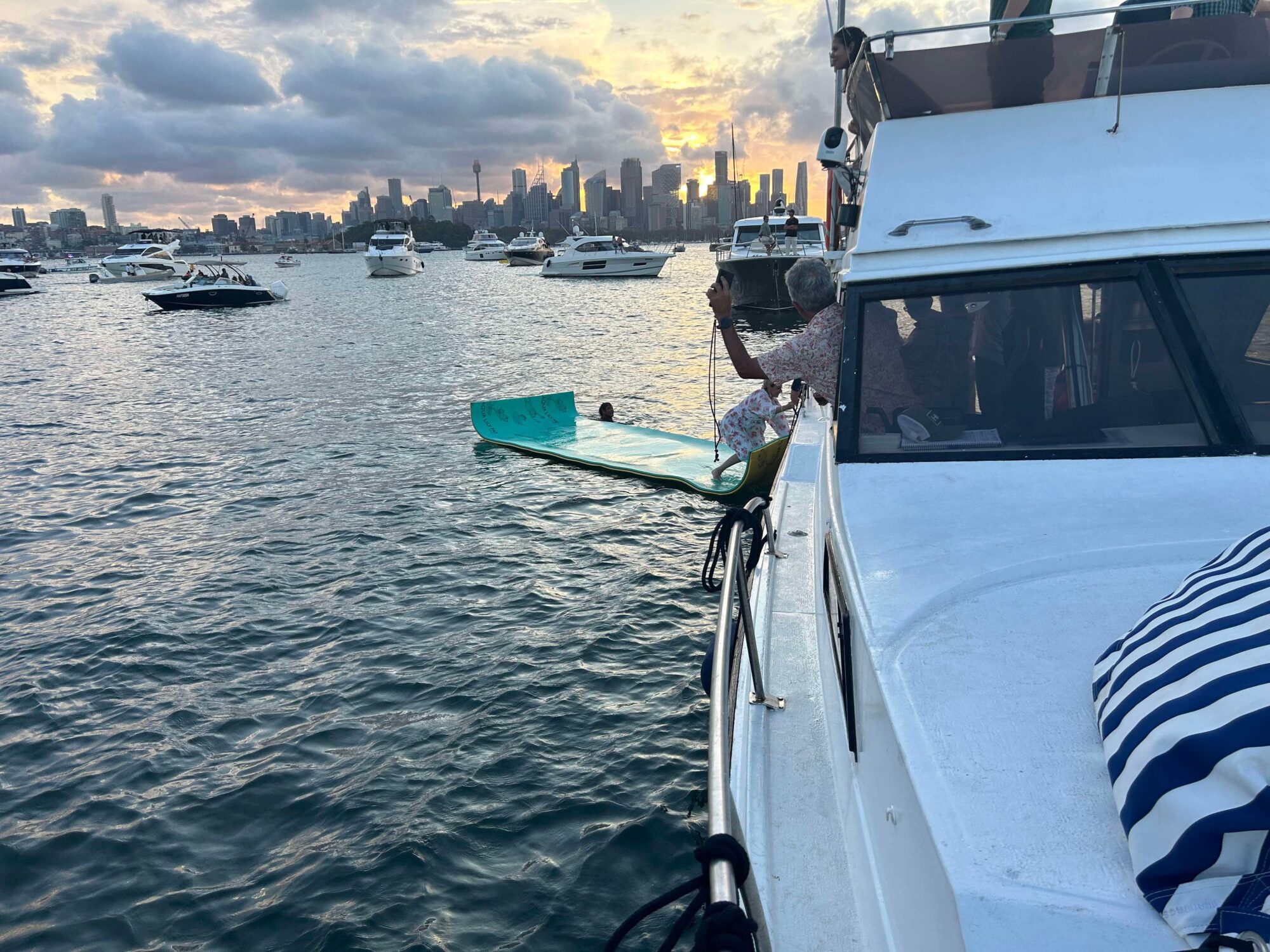 A boat at sunset shows a woman adjusting a green Mayfair float in the water, with anchored boats nearby and a city skyline under a cloudy sky.