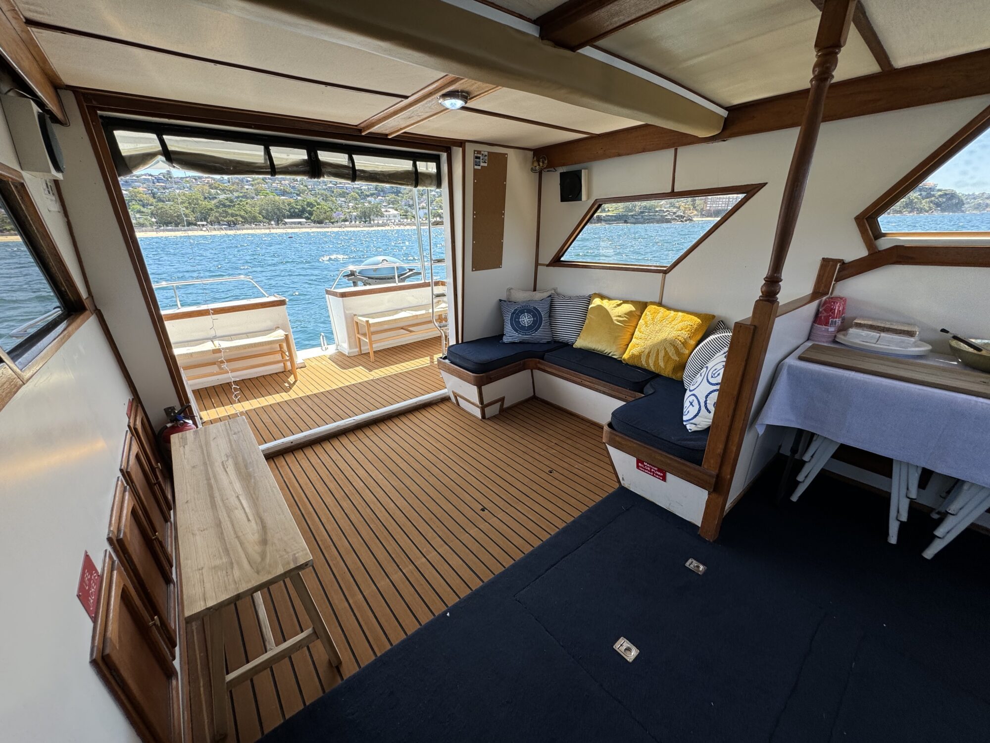 The Mayfair features a boat interior with wooden flooring, cushioned corner seating with pillows, a table, and large windows. A door leads to the deck for views of blue water and a scenic shoreline.