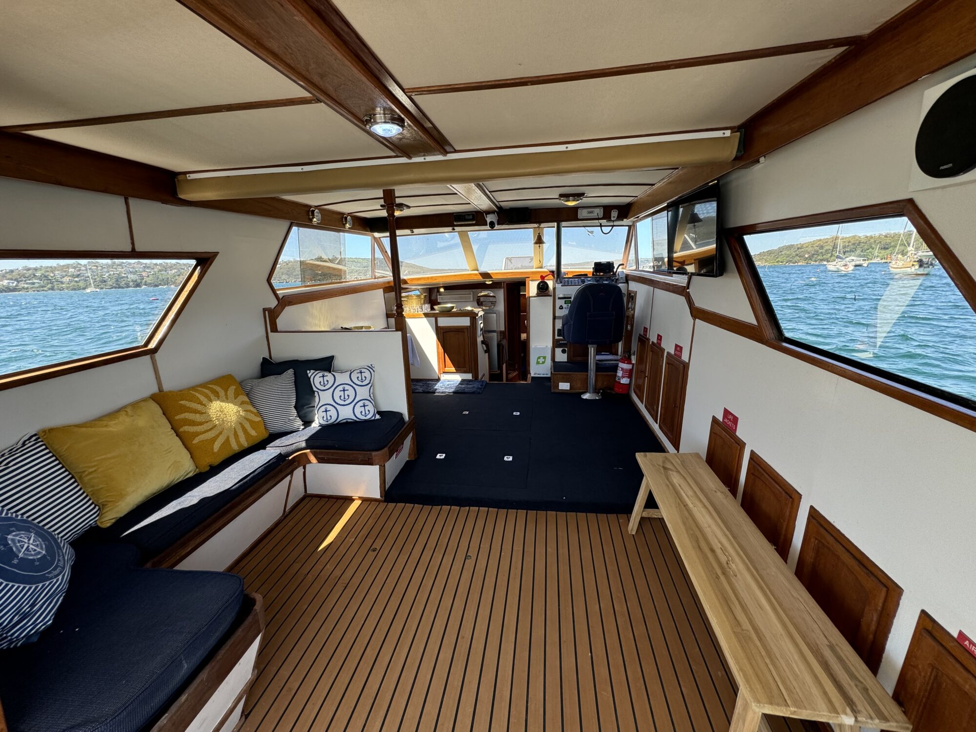 The Mayfair features a wooden interior with cushioned seating, a bench, and large windows offering blue water views and a distant shoreline under clear skies. The helm is visible at the front.