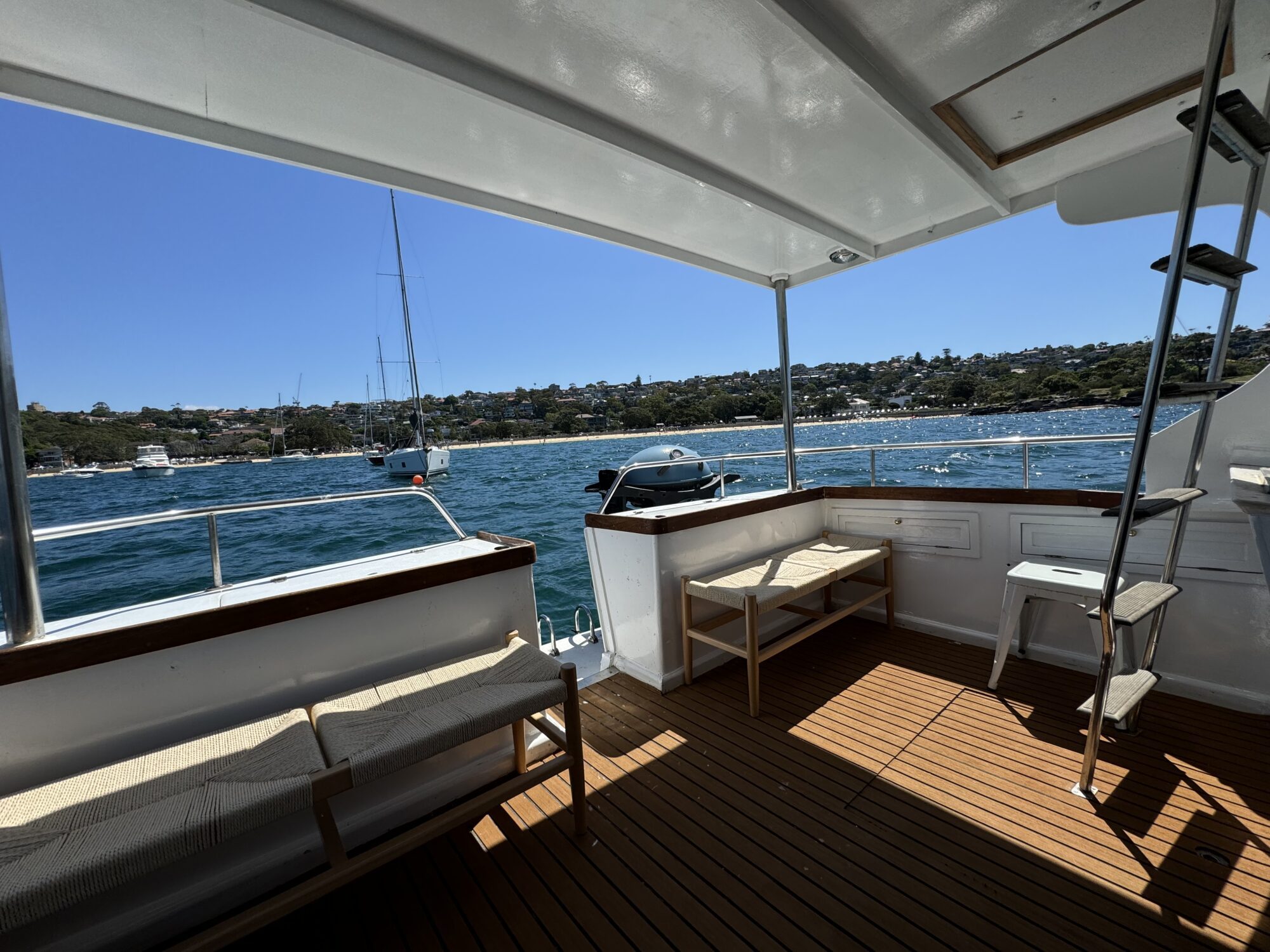 View from the covered deck of Mayfair, featuring wooden benches and a chair. The boat floats on blue water near a tree-lined coast with houses under a clear sky. A sailboat is seen in the distance.