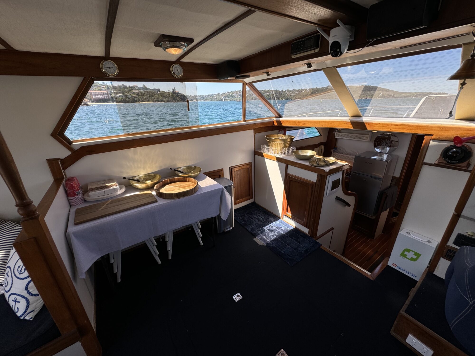 The interior of the Mayfair features a dining table with plates and bowls on a white tablecloth, wood paneling, windows displaying water and land, and a compact kitchenette area to the right.