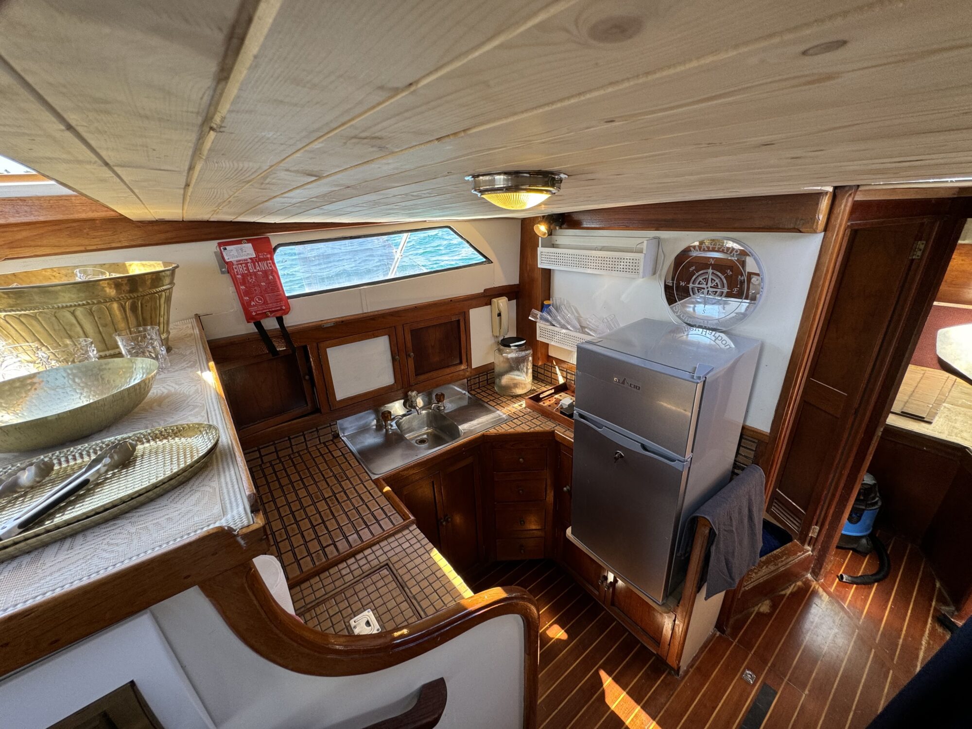 The Mayfair features a cozy, nautical boat kitchen with wood paneling, sink, countertop, refrigerator, microwave, and accessories. Sunlight from the window above the sink enhances the inviting interior.