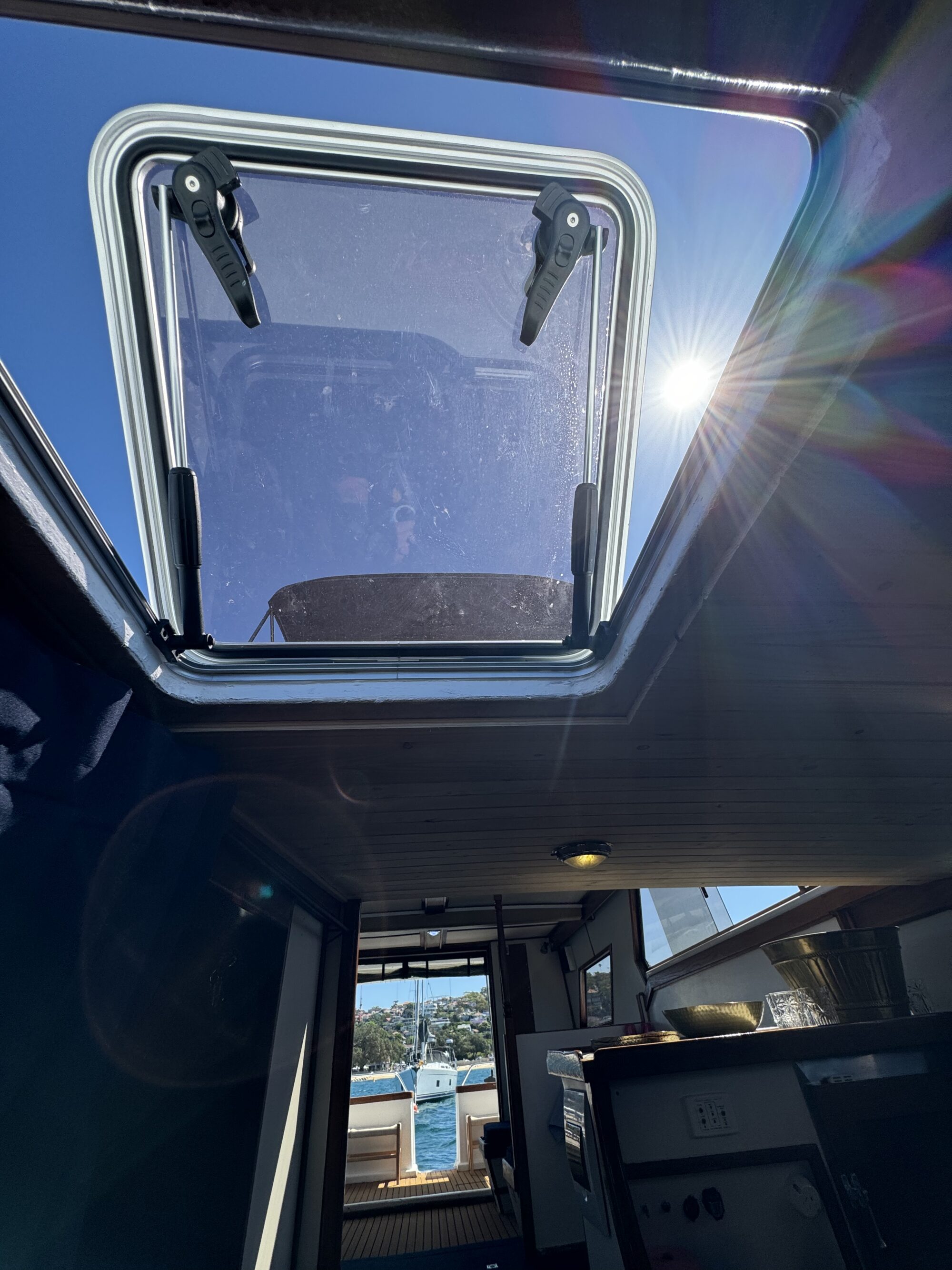 View from inside a Mayfair cabin looking up through an open sunroof, sunlight streaming in, with blue sky and the docked marina visible through the open rear door.