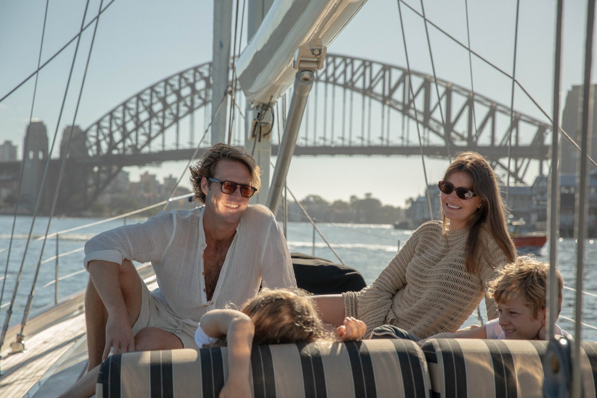 A smiling family of four relaxes on a sailboat in sunny weather, with a large bridge and city skyline in the background. The adults wear sunglasses and light clothing as they lounge together.