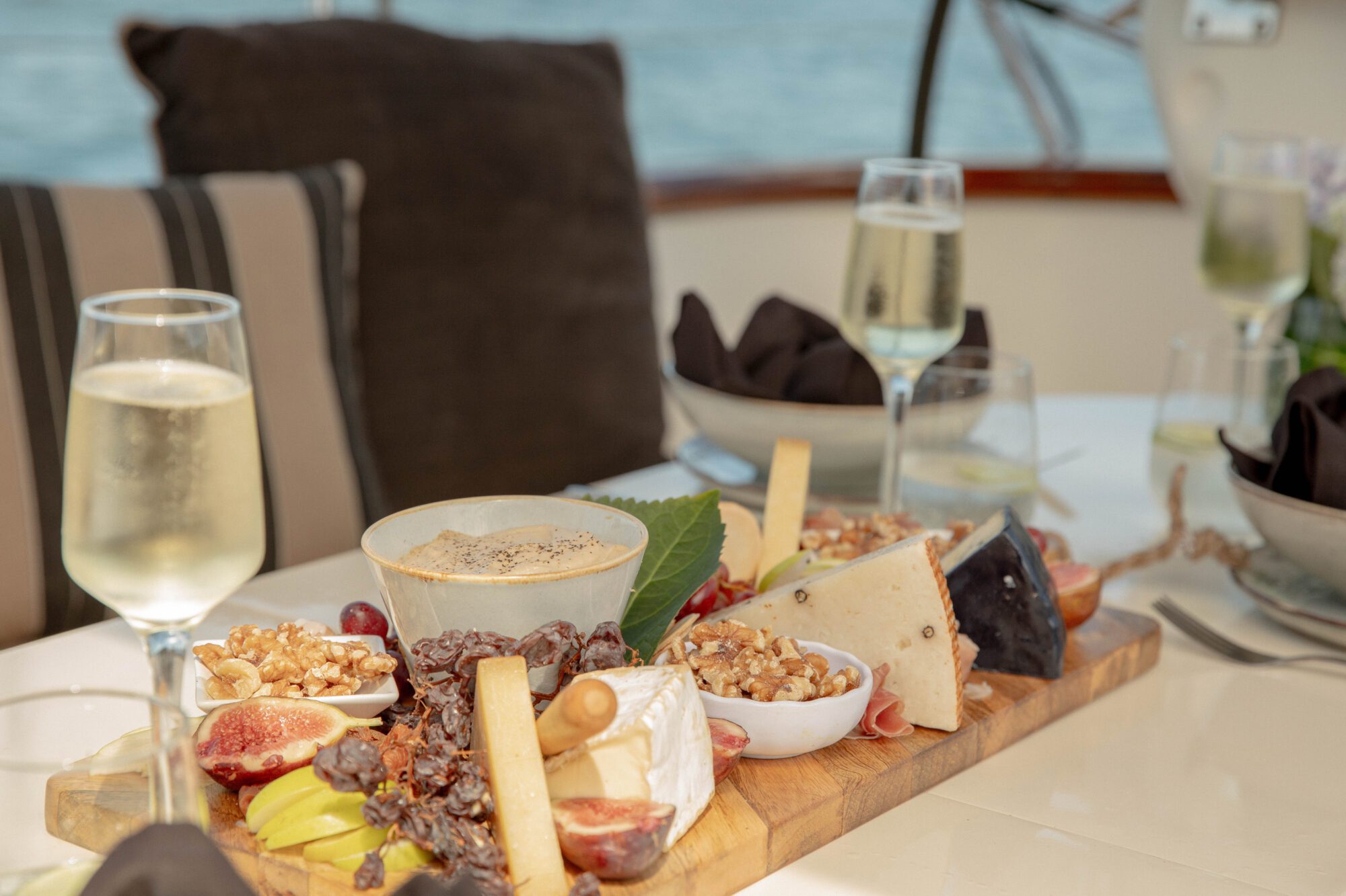 A wooden board with assorted cheeses, nuts, figs, grapes, and breadsticks sits on a table next to two glasses of white wine, with a blurred background of a boat and the sea.