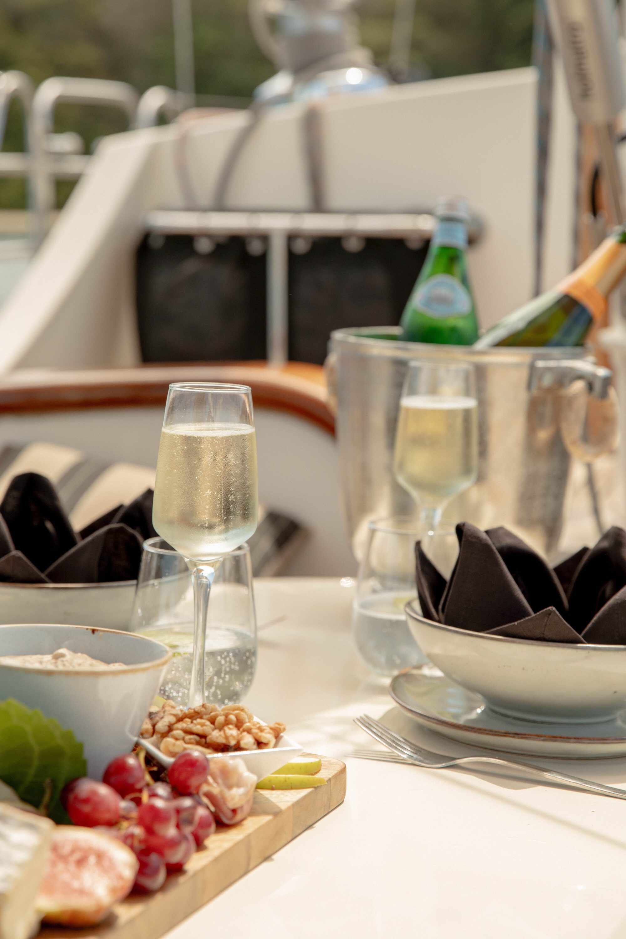 A glass of champagne on a table set with food, napkins, and drinks on a boat deck, with a charcuterie board featuring grapes, nuts, and cheese in the foreground.