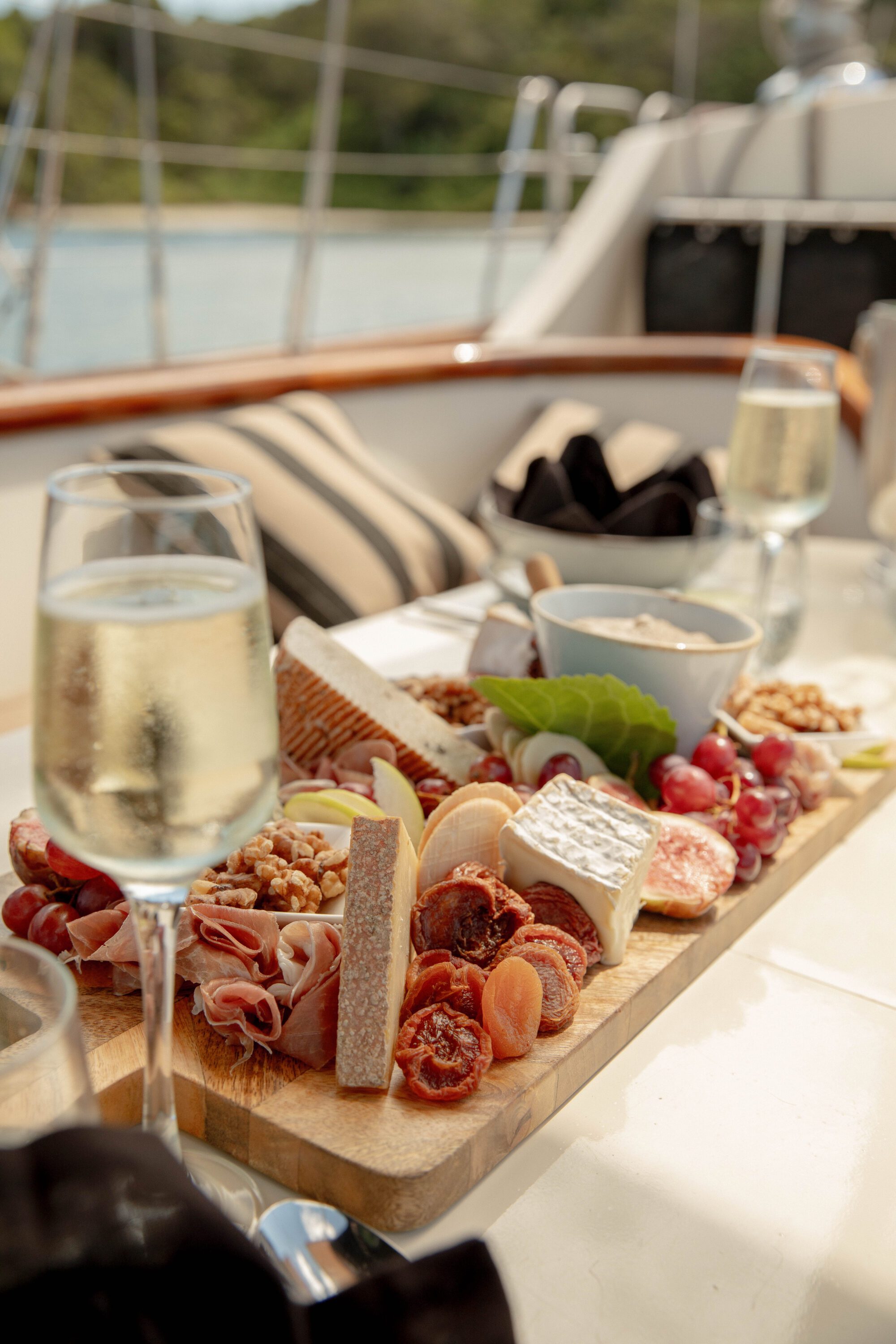 A wooden board on a yacht table holds assorted cheeses, cured meats, dried tomatoes, grapes, nuts, and crackers. Two glasses of sparkling wine are beside the board. The background shows a blurred water and greenery view.