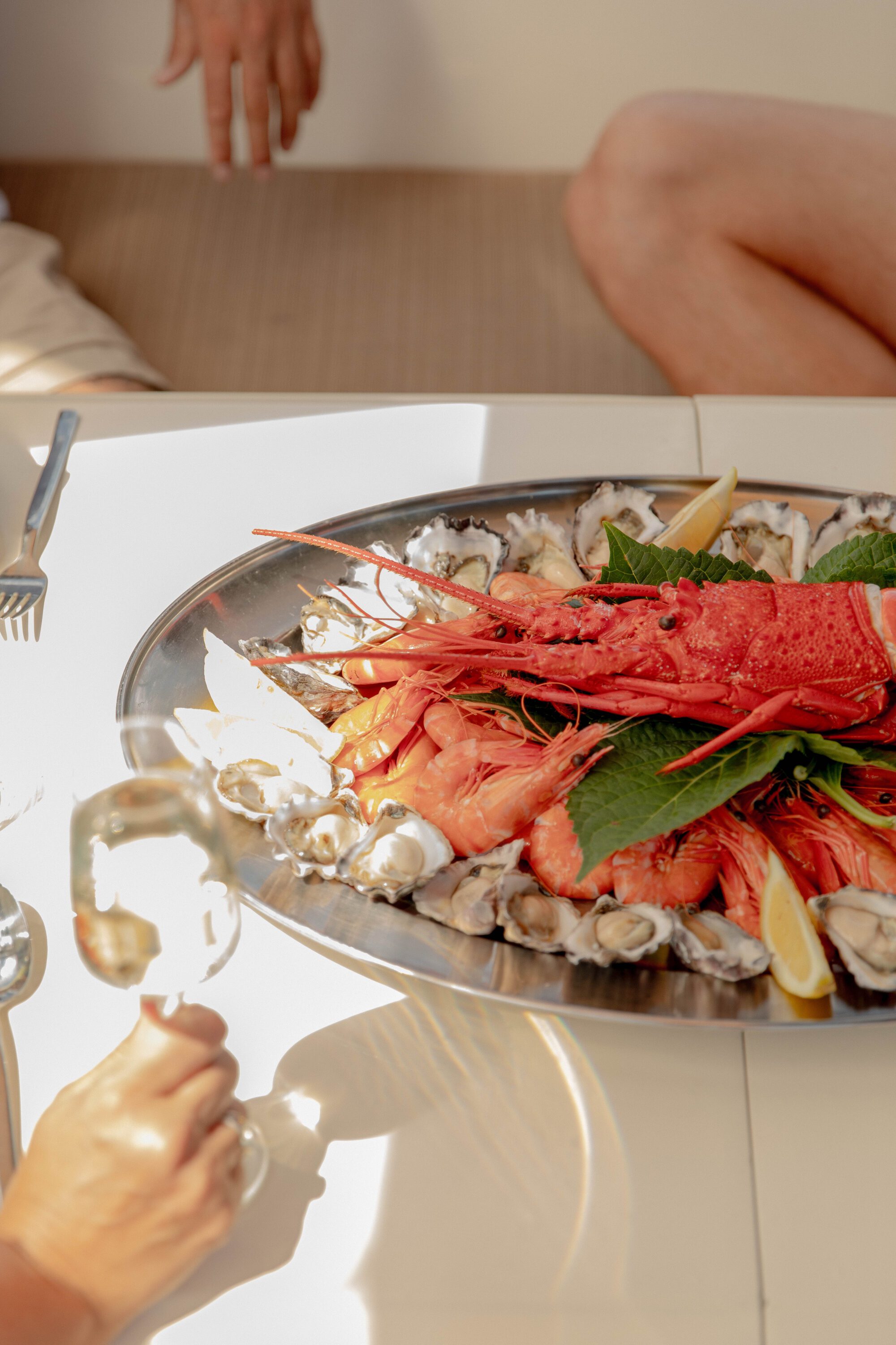 A seafood platter with lobster, shrimp, oysters, lemon wedges, and greens sits on a table. A person holds a glass of wine, and two others are partially visible sitting nearby.
