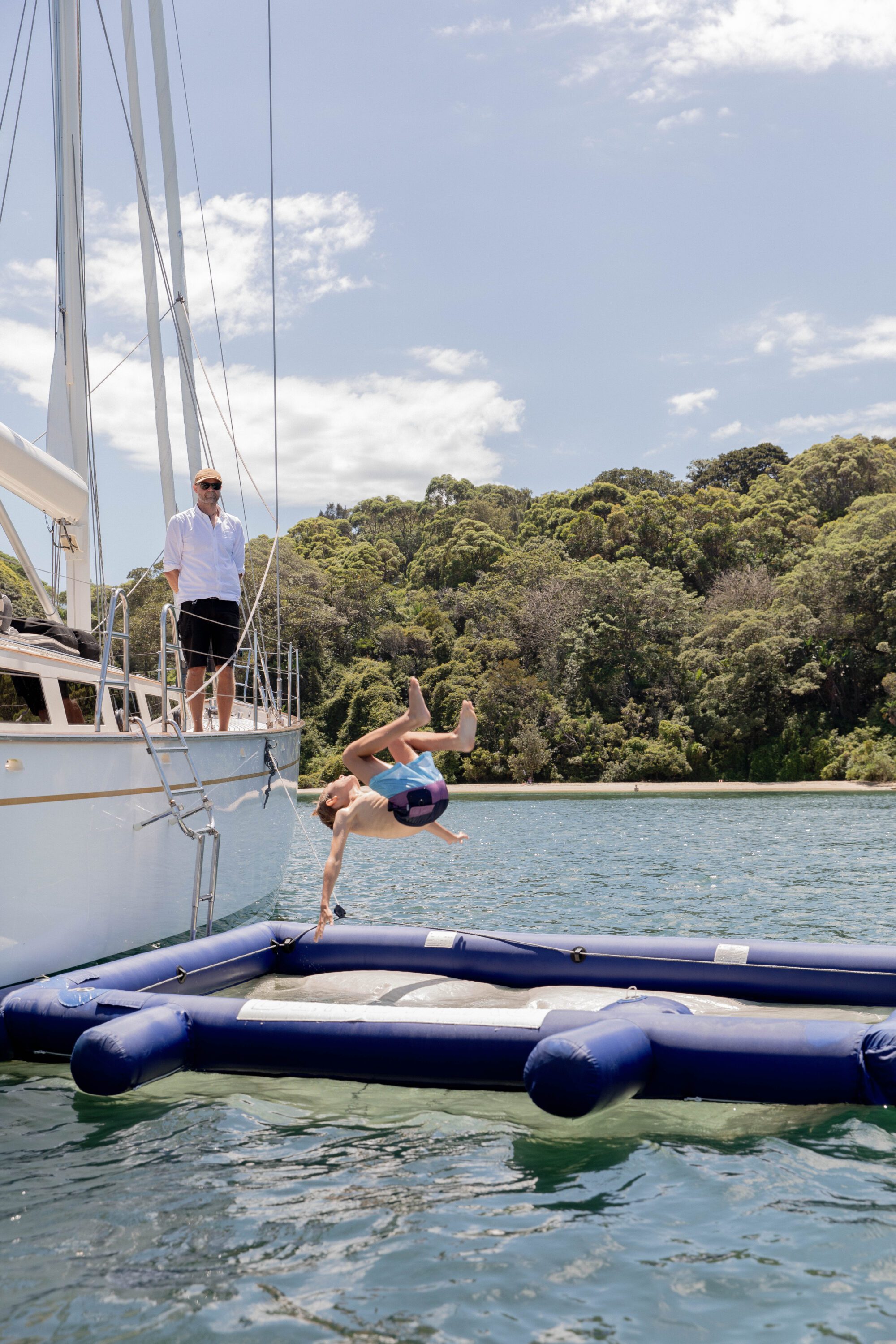 A person in swim trunks is mid-flip off a floating inflatable dock next to a sailboat, while another person stands on the boat, watching. Trees and blue sky are visible in the background.