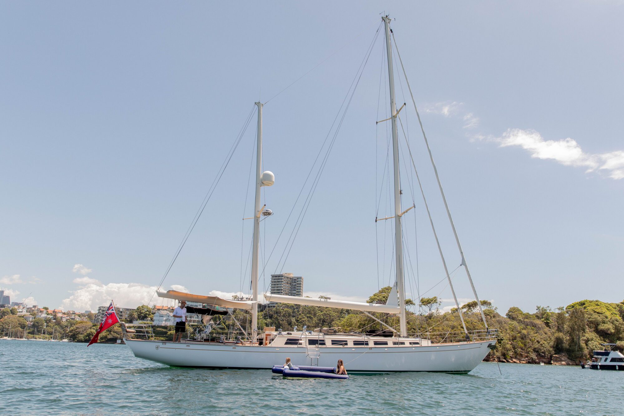 A large white sailboat is anchored on calm water near a green shoreline, with two people sitting in a small inflatable boat nearby. Buildings are visible in the background under a partly cloudy sky.