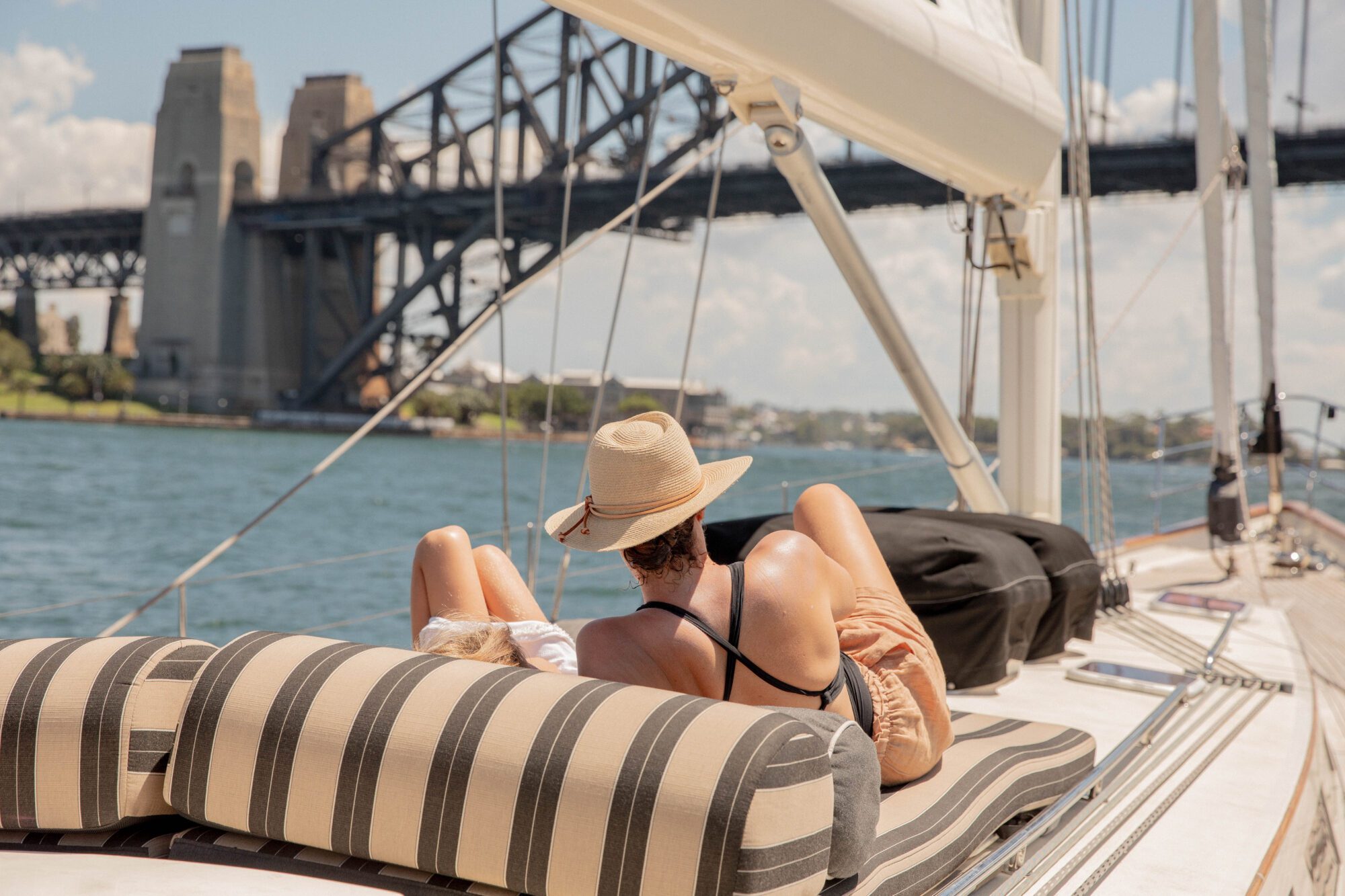 A person wearing a hat relaxes on a striped lounge chair on a boat, with the Sydney Harbour Bridge and waterfront visible in the background under a sunny sky.