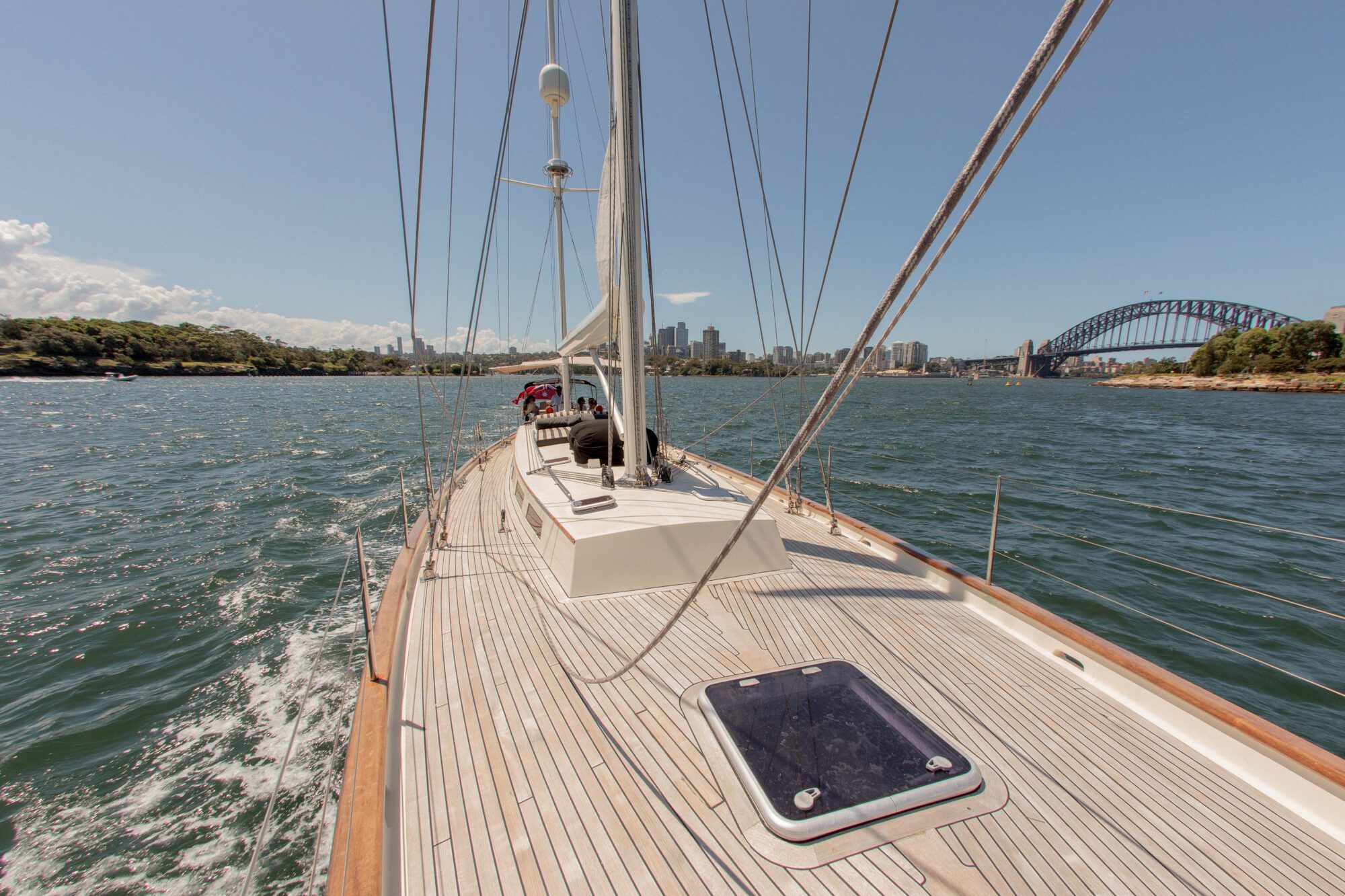 View from the deck of a sailboat on the water, heading towards a city skyline with tall buildings and a large arch bridge in the background under a clear blue sky.