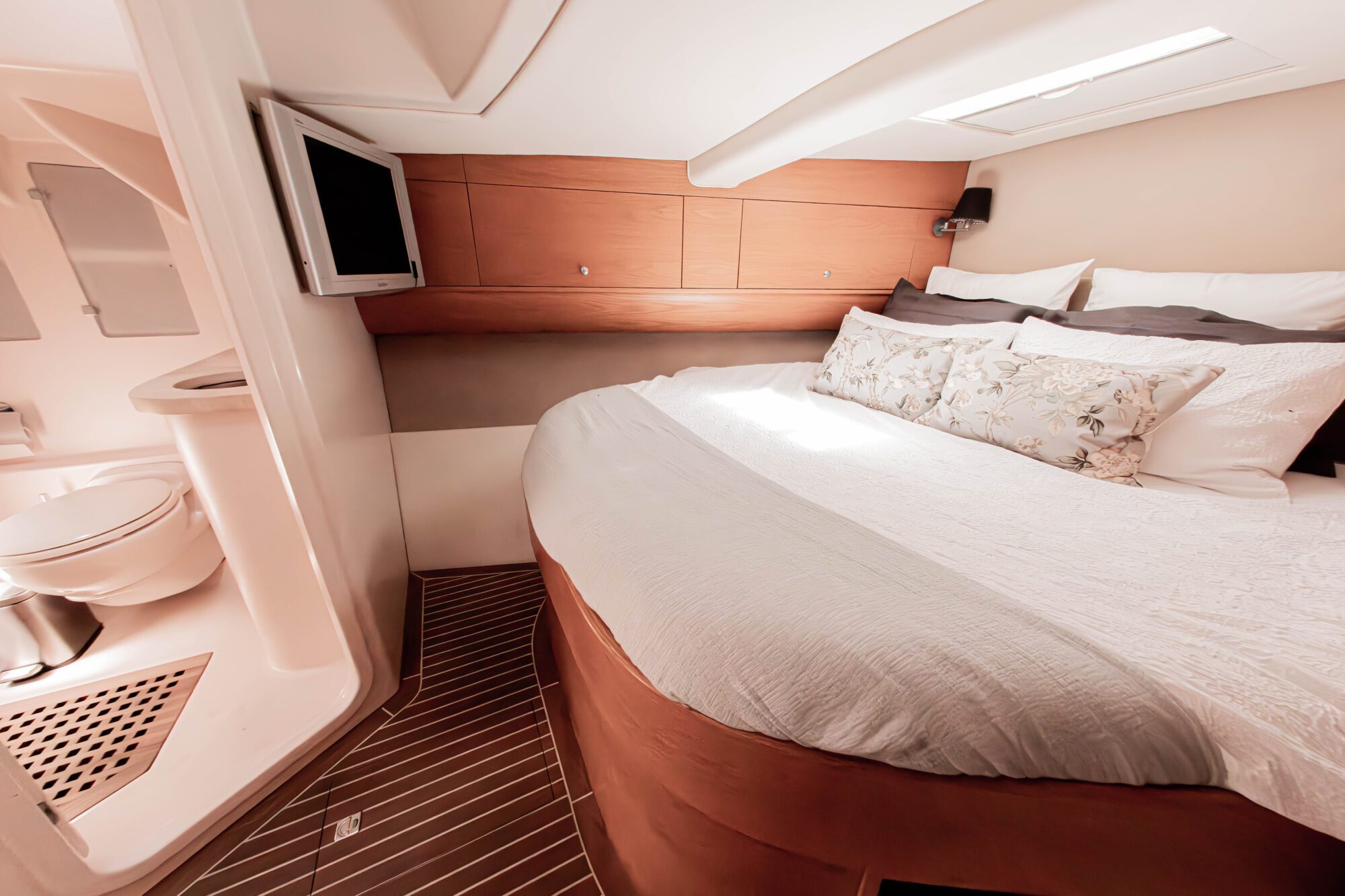 A cozy cabin bedroom on a boat features a rounded bed with white and floral pillows, wood paneling, a wall-mounted TV, and an adjacent compact bathroom with a toilet and shower. Natural light enters from a ceiling window.