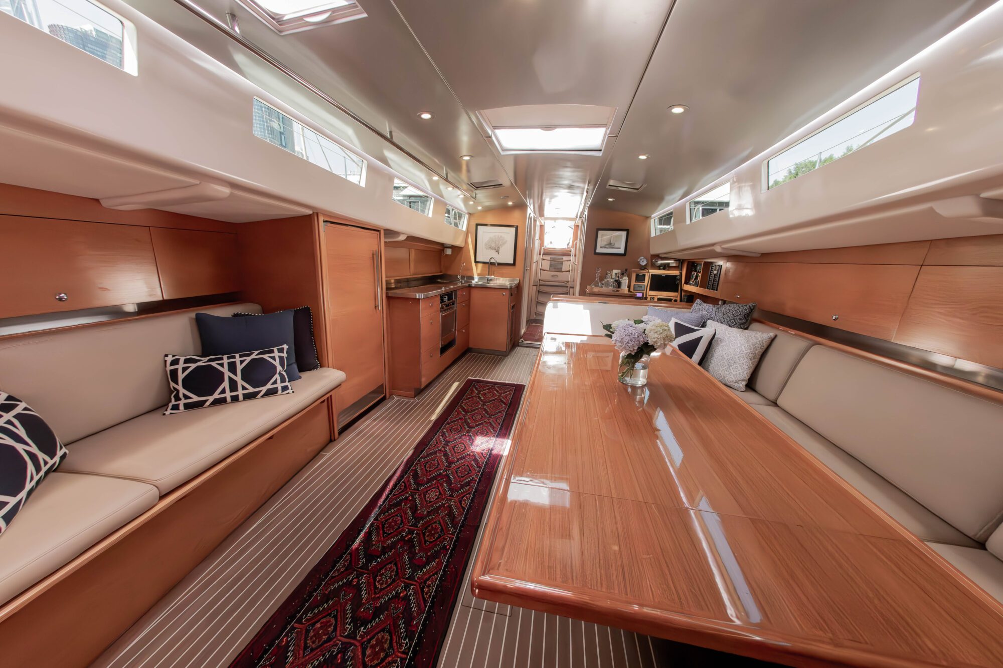 The interior of a yacht featuring wooden paneling, a long polished dining table with flowers, cushioned seating with pillows, a kitchen area, and natural light from skylights above.