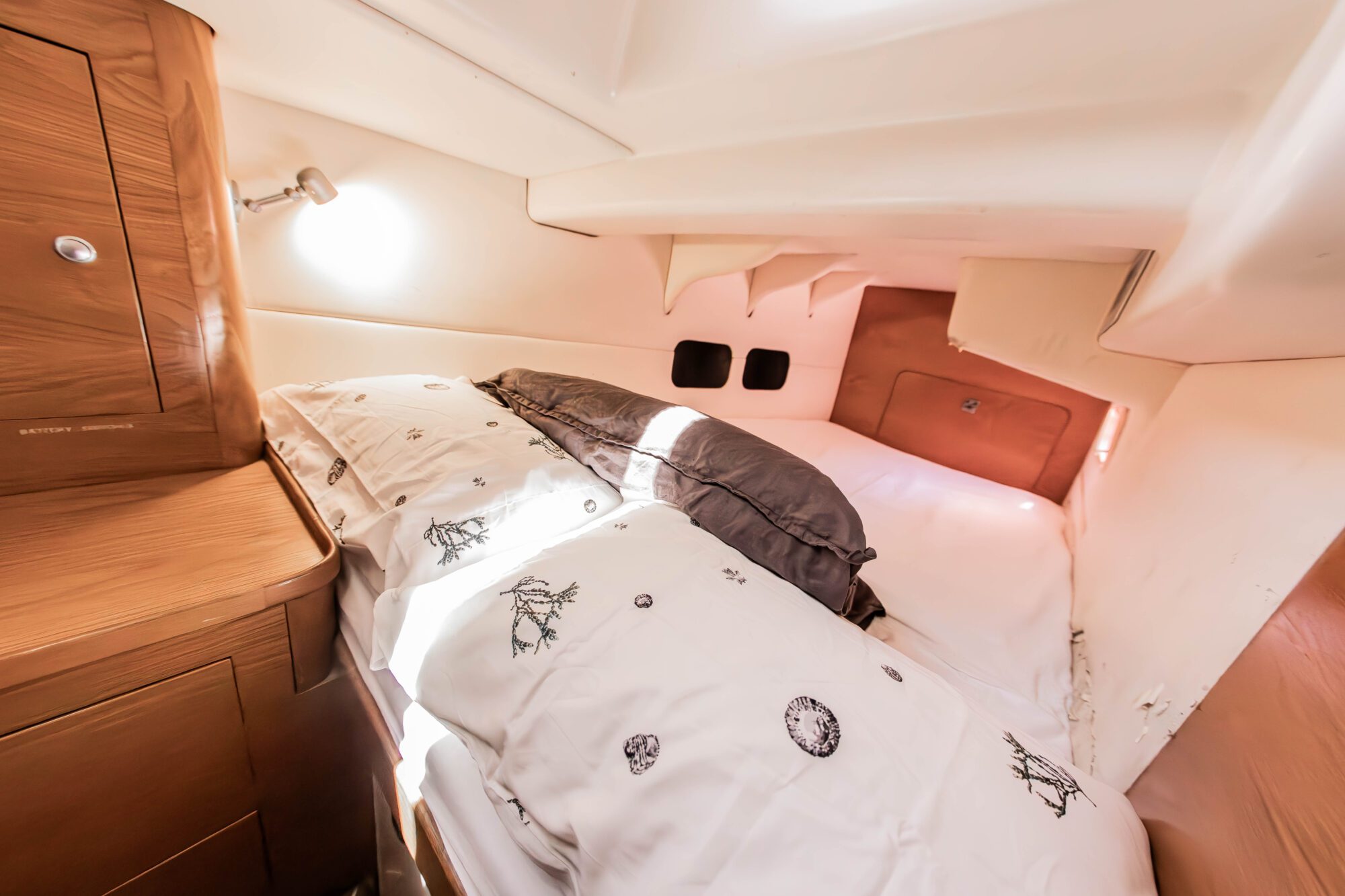 A cozy boat cabin with a double bed, white bedding with black sea-themed prints, a brown blanket, wooden cabinets, a small wall lamp, and two porthole windows letting in natural light.