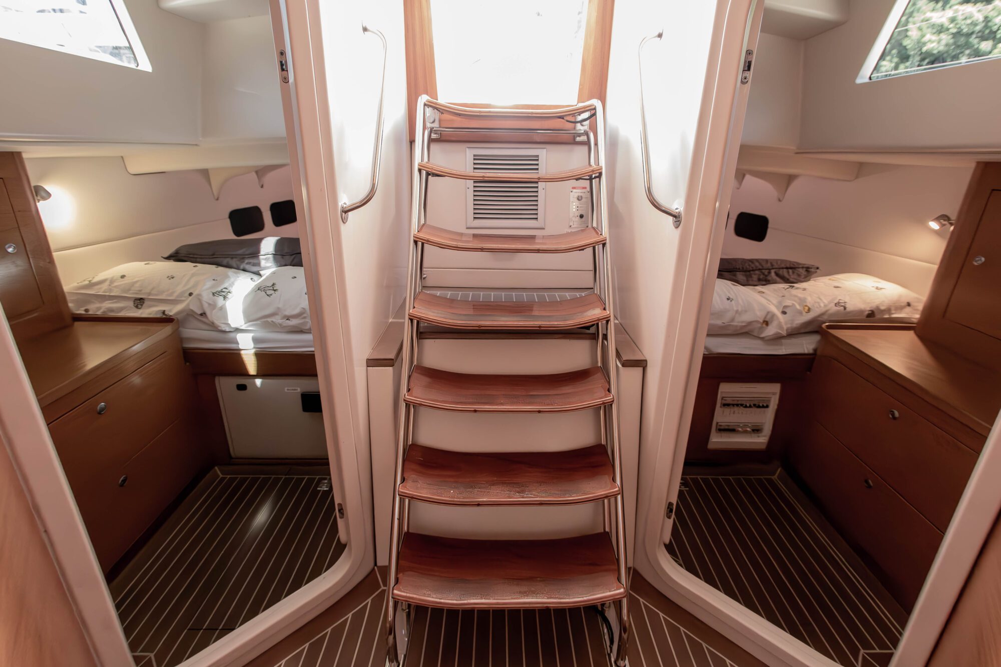 Wooden steps lead down into a boat’s cabin, flanked by two cozy bedrooms with beds and wooden storage. Soft natural light comes from porthole windows above each bed, and the floor has striped wood paneling.