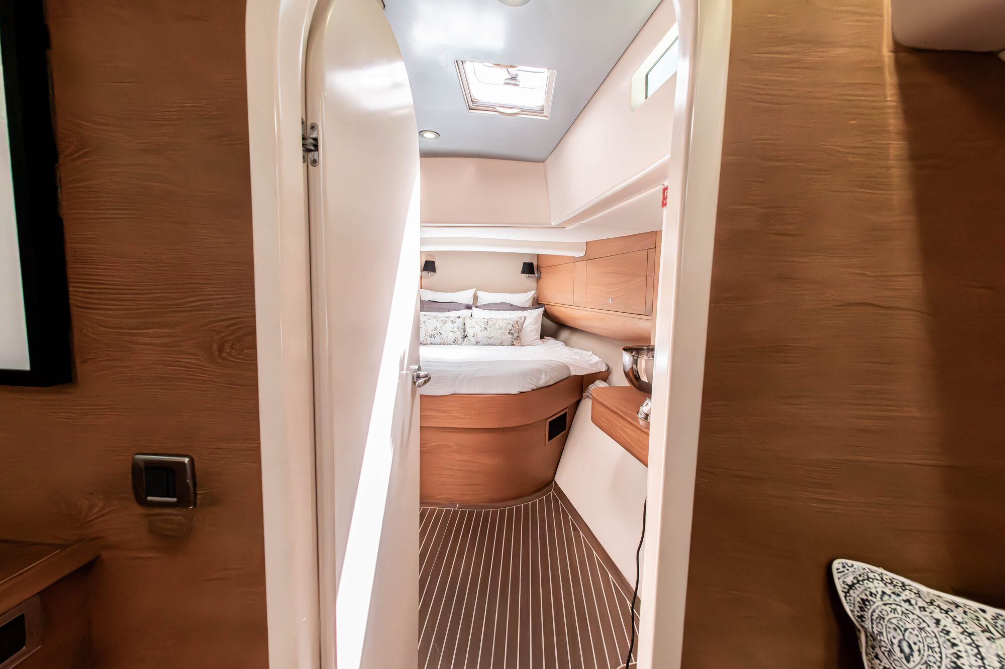 View through a partially open door into a cozy boat cabin with a double bed, decorative pillows, wood paneling, striped flooring, and a skylight above.