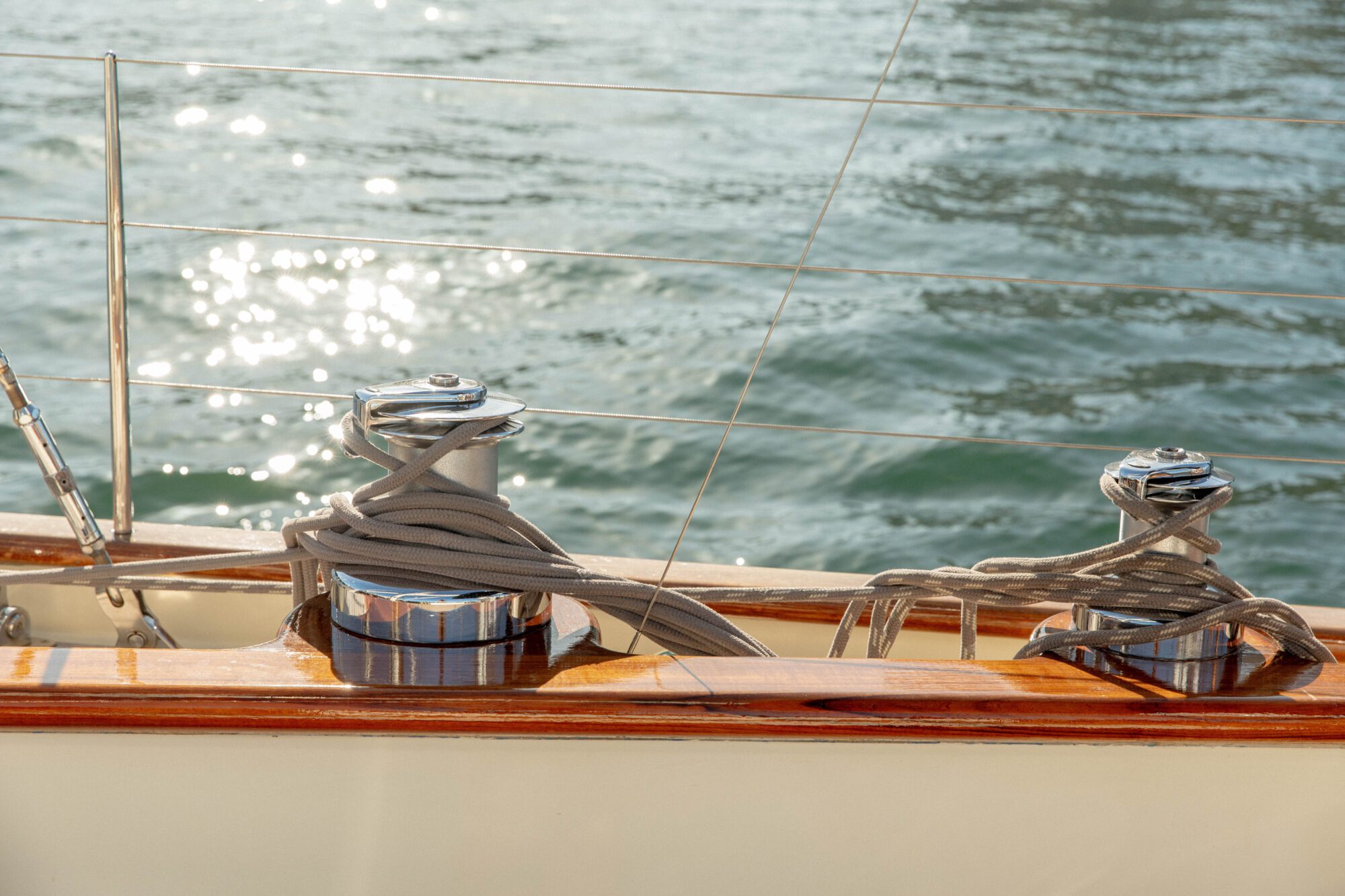 Close-up of coiled ropes on shiny metal cleats on a wooden sailboat deck, with sunlight reflecting off the water in the background.