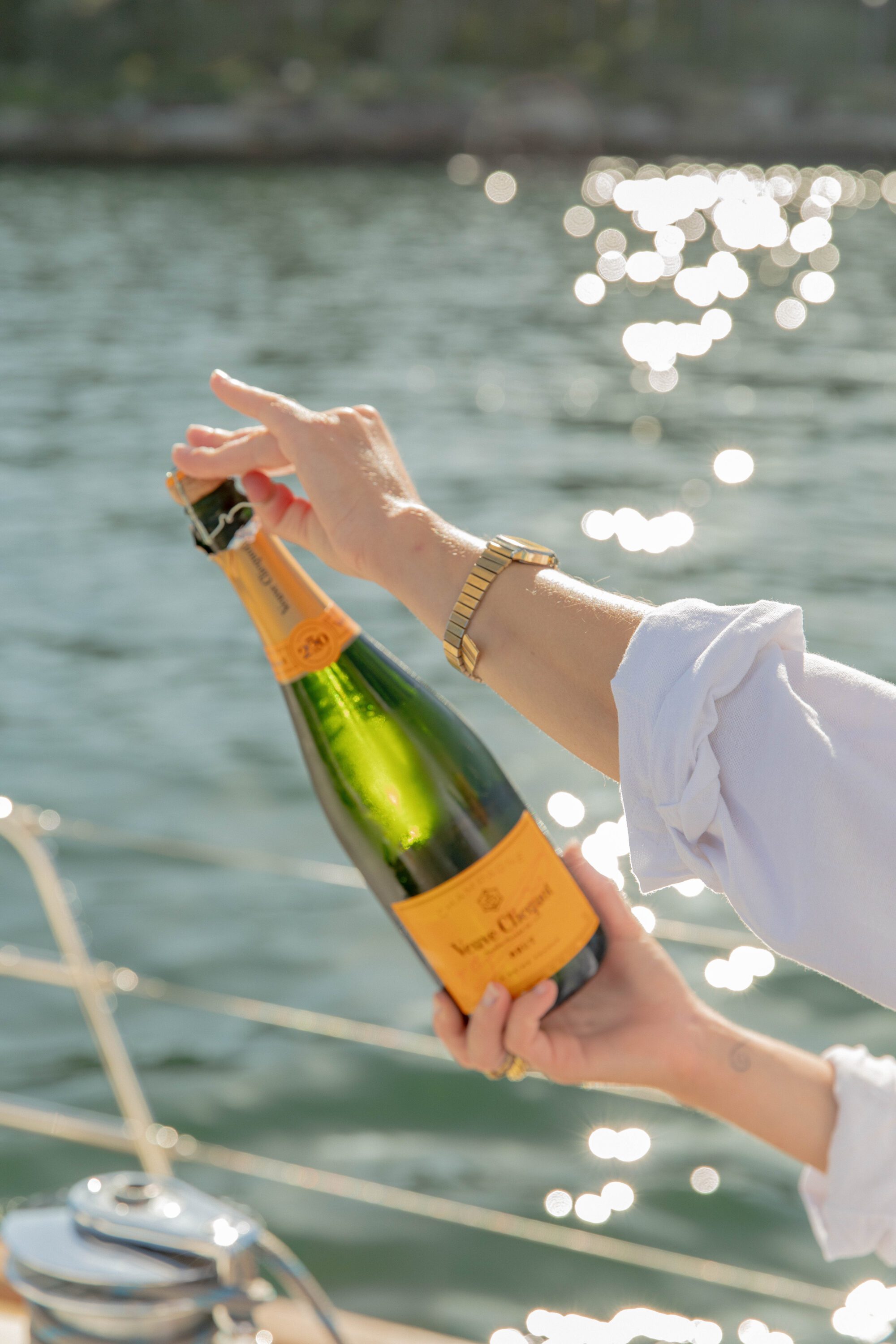 A person in a white shirt opens a bottle of Veuve Clicquot champagne on a boat, with sparkling water in the background.